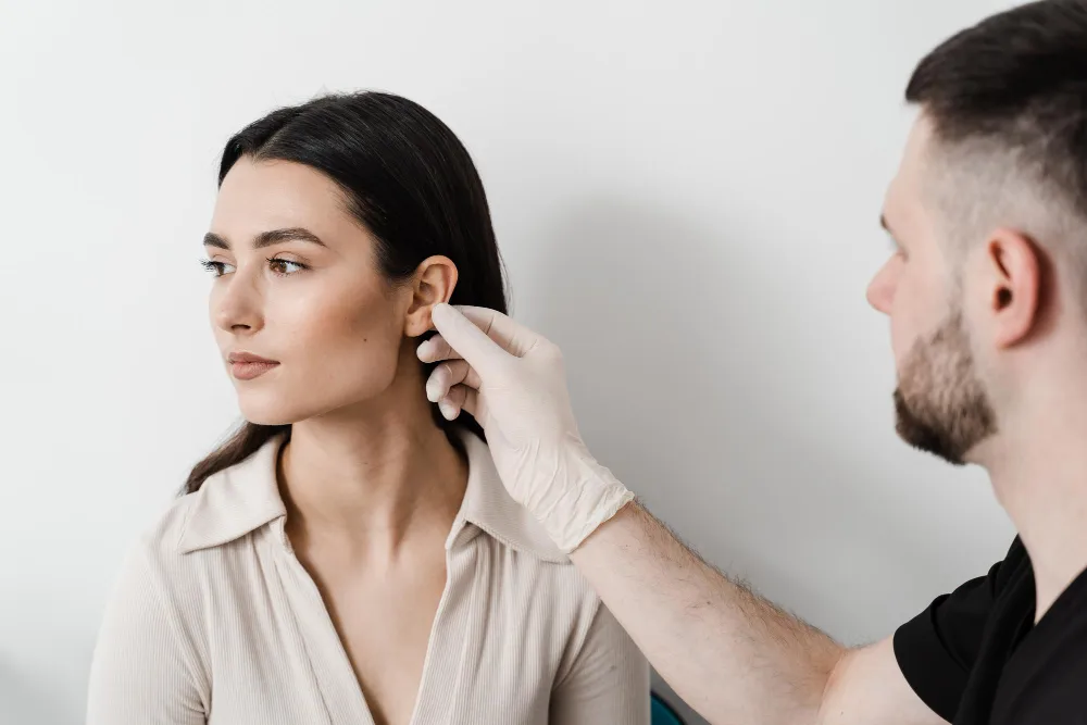 Man wearing gloves examines a woman's ear in a medical or clinical setting with white background.