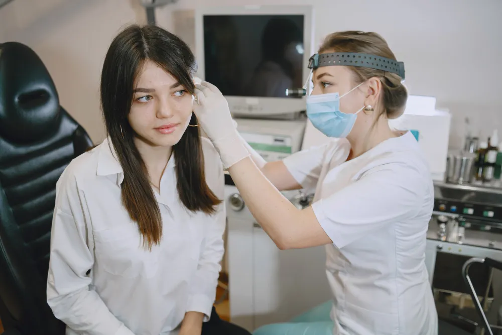 A healthcare professional wearing mask and gloves examines a young woman's ear in a clinic.
