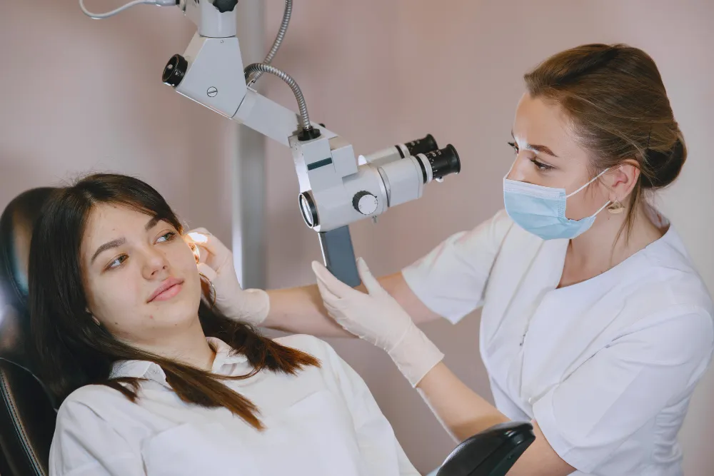 Healthcare professional wearing mask examining patient's ear with otoscope and microscope in clinic.