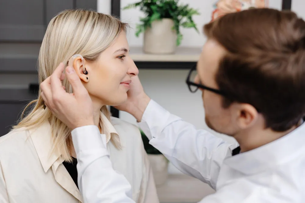A man fitting a hearing aid on a smiling blonde woman in a clinical setting.