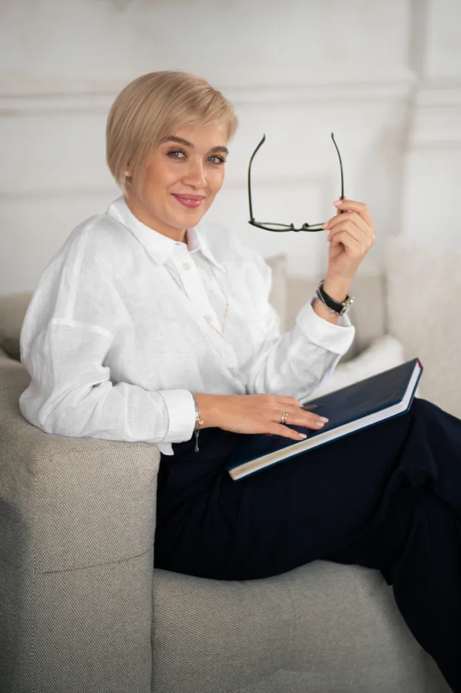 Smiling woman sitting on sofa holding glasses in one hand and a closed book in her lap.