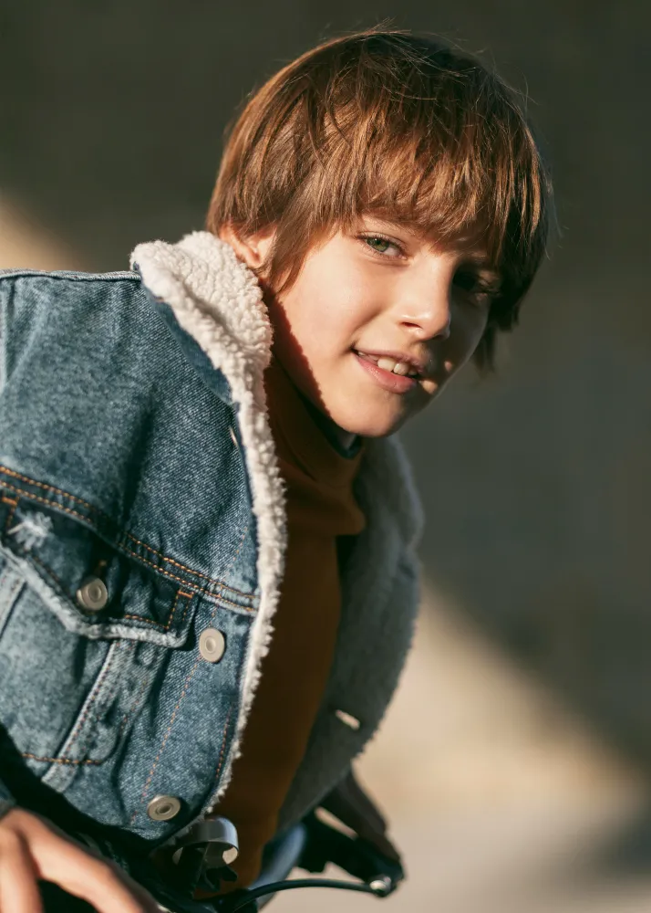 Young boy wearing a denim jacket with shearling lining, smiling and leaning forward outdoors.