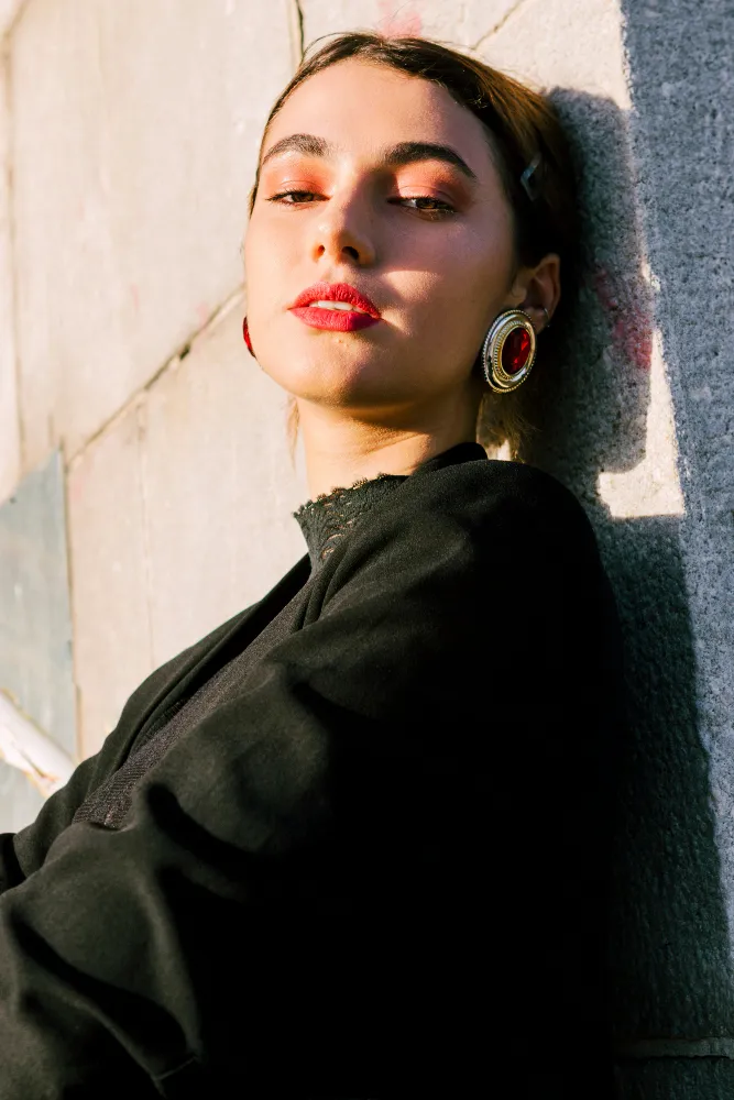 Young woman with red lipstick and large red earrings leaning against a sunlit stone wall.