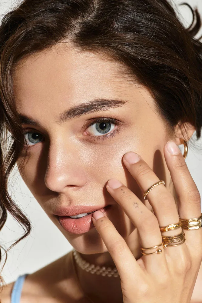 Close-up of a woman with blue eyes touching her face, wearing multiple gold rings and a pearl choker.