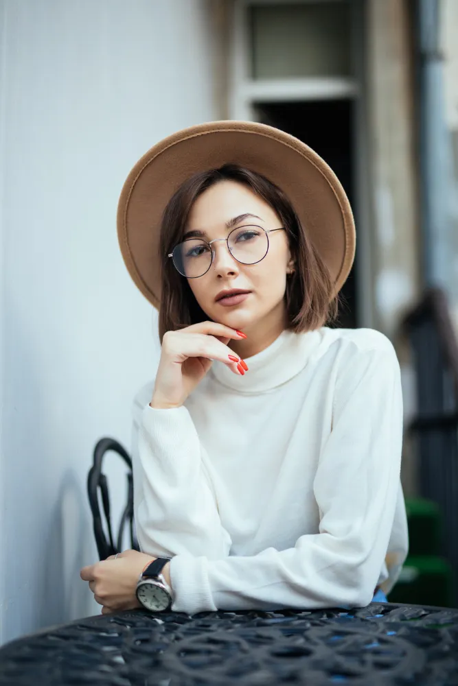 Young woman with round glasses and beige hat sitting at a table, resting her chin on her hand.