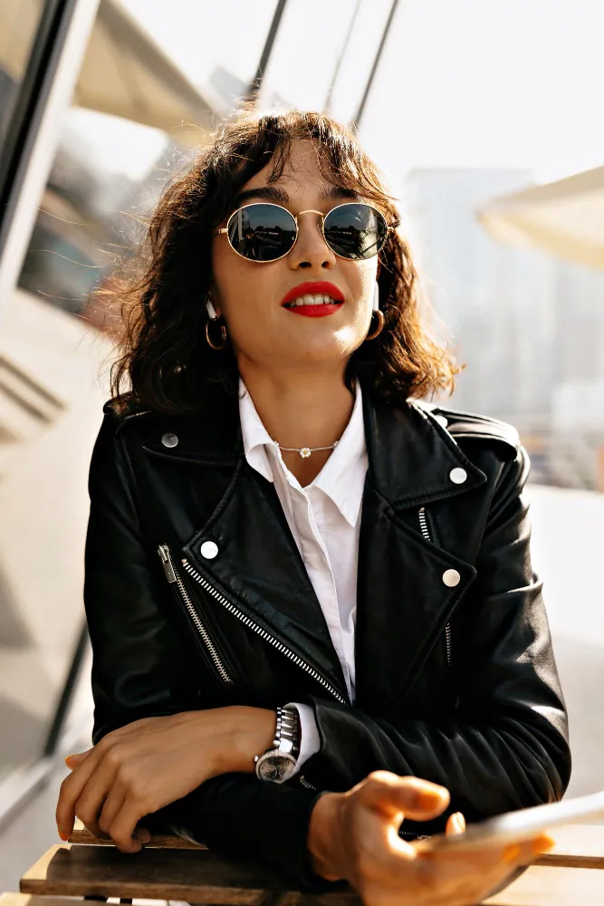 Woman with short curly hair wearing sunglasses and a black leather jacket, sitting at a table outdoors.