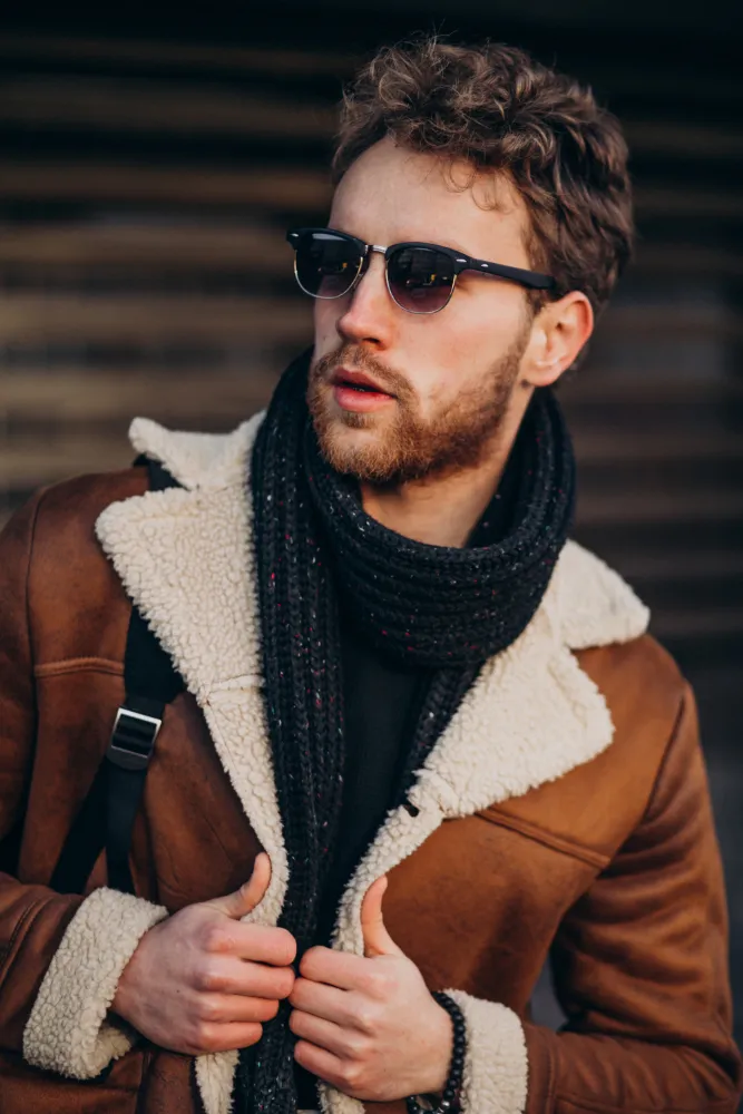 Man with curly hair wearing sunglasses, a brown shearling jacket, and a black scarf, looking sideways.