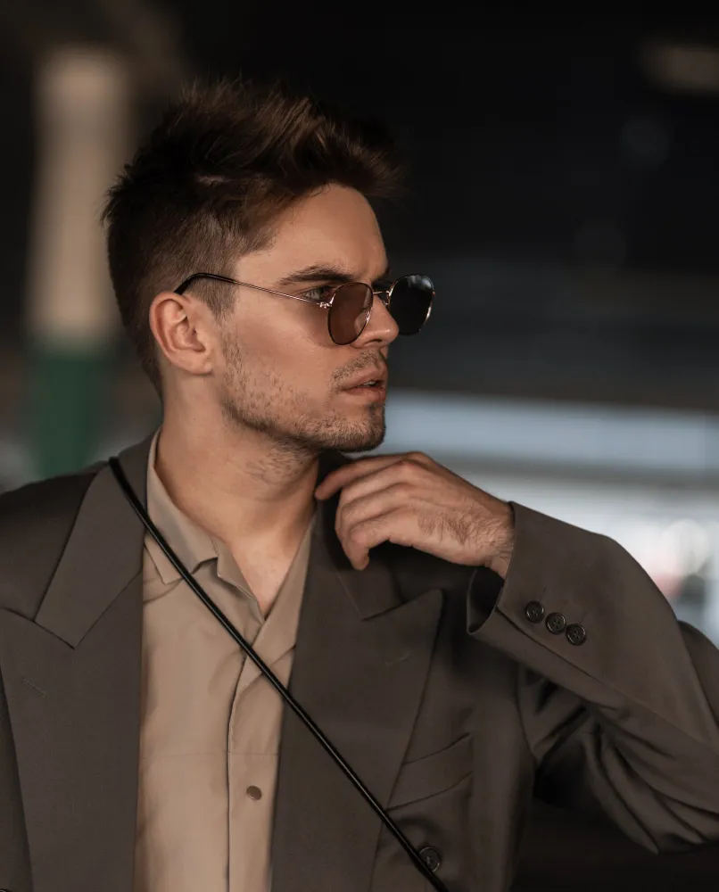 Man with sunglasses and spiked hair wearing a dark blazer and beige shirt, looking to the side.