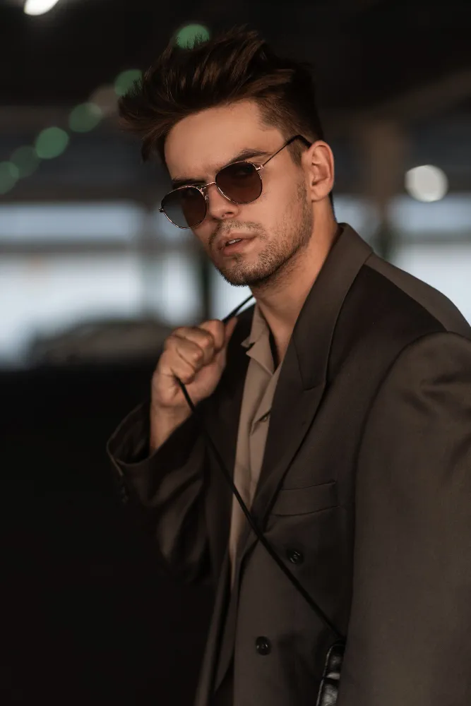 Man wearing sunglasses and a dark blazer holding a strap over his shoulder with blurred lights behind.