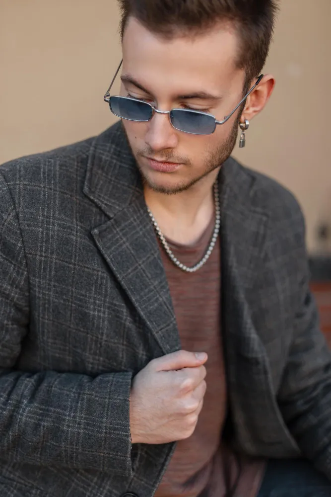 Young man wearing blue sunglasses, a plaid blazer, and a chain necklace looking down.