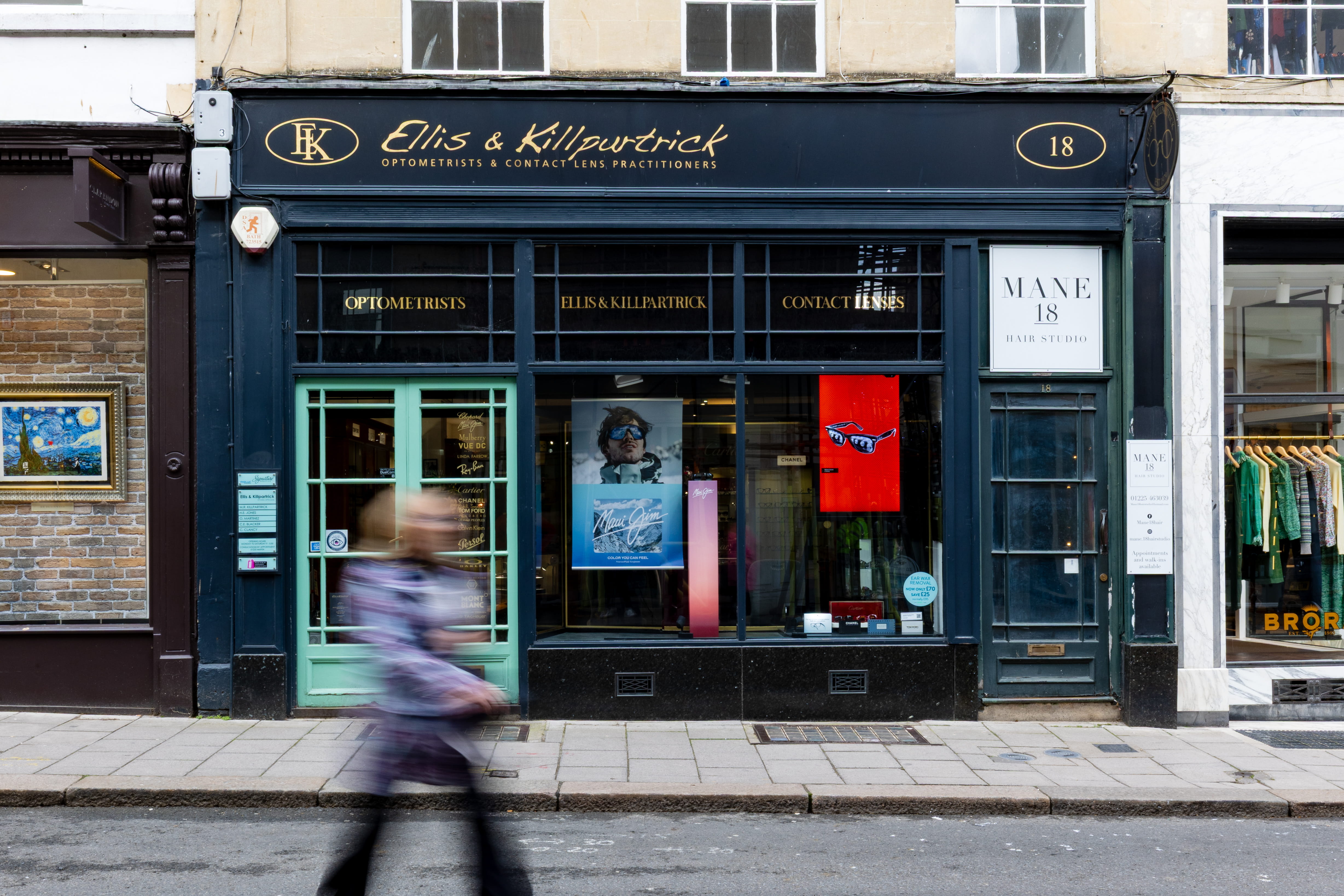 Storefront of Ellis & Killpartrick optometrists with black sign and green door, a blurred person walking by.