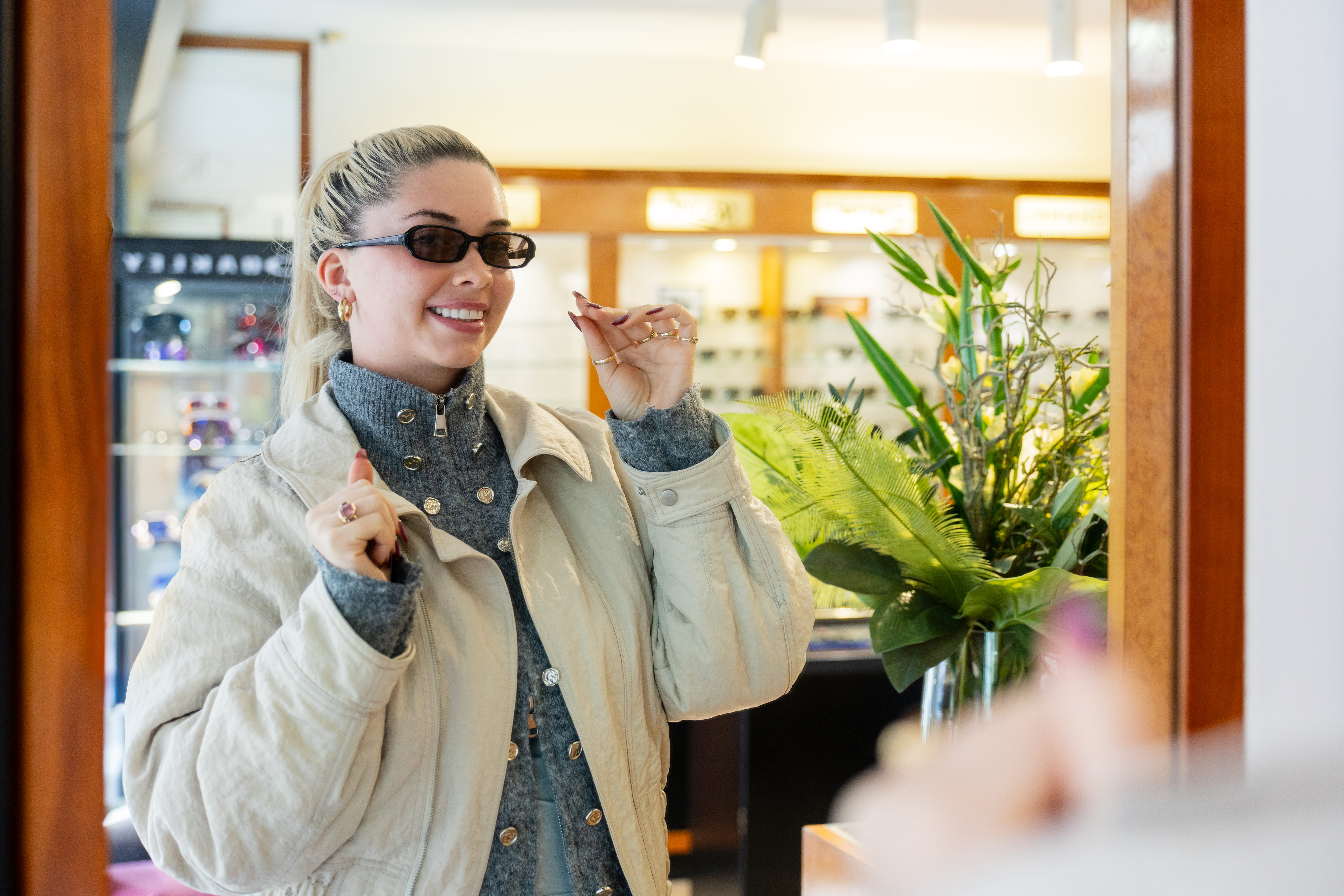 Smiling woman wearing sunglasses and a beige jacket looking at herself in a mirror inside a store.