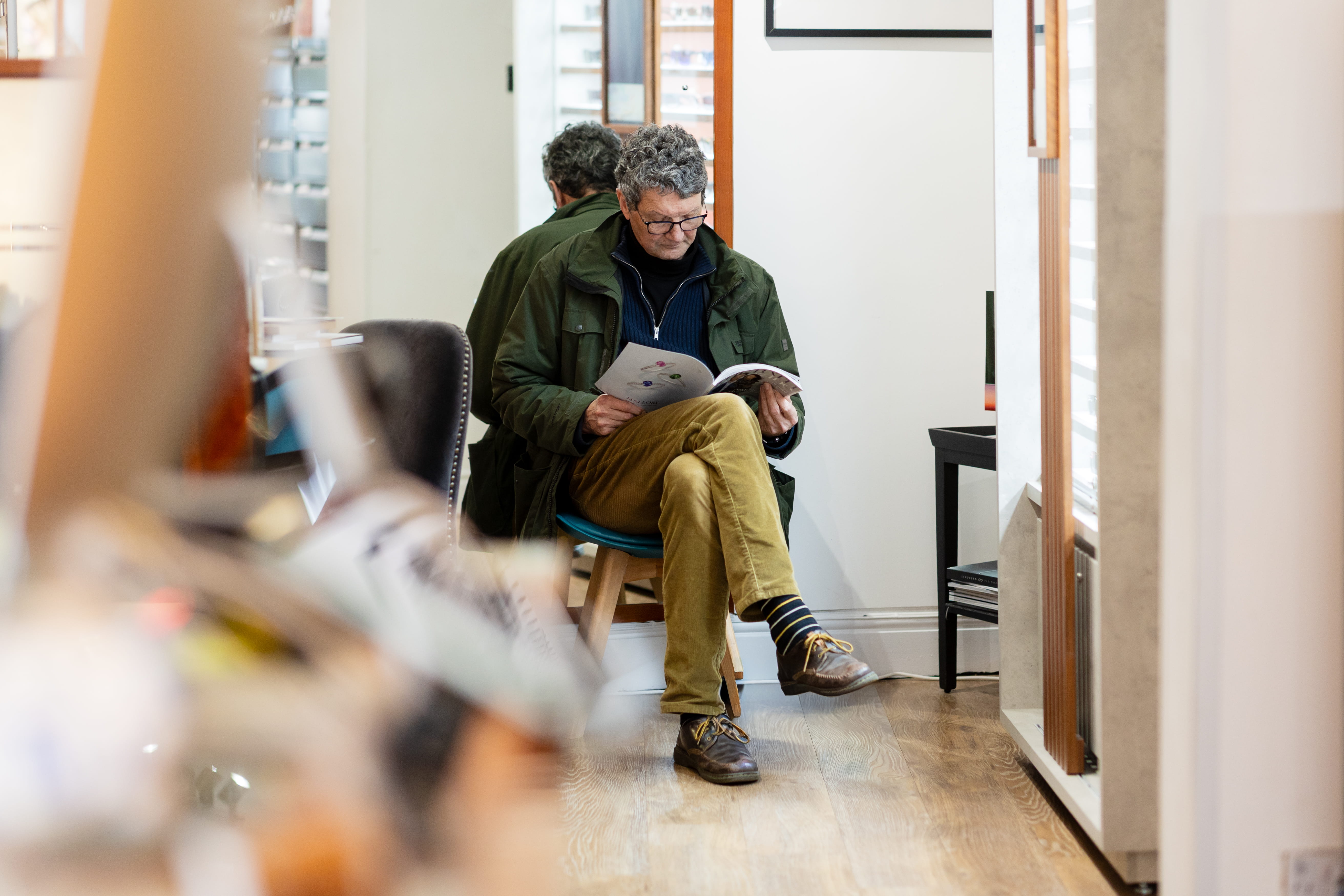 Man with glasses in green jacket and striped socks sitting on stool reading a magazine indoors.
