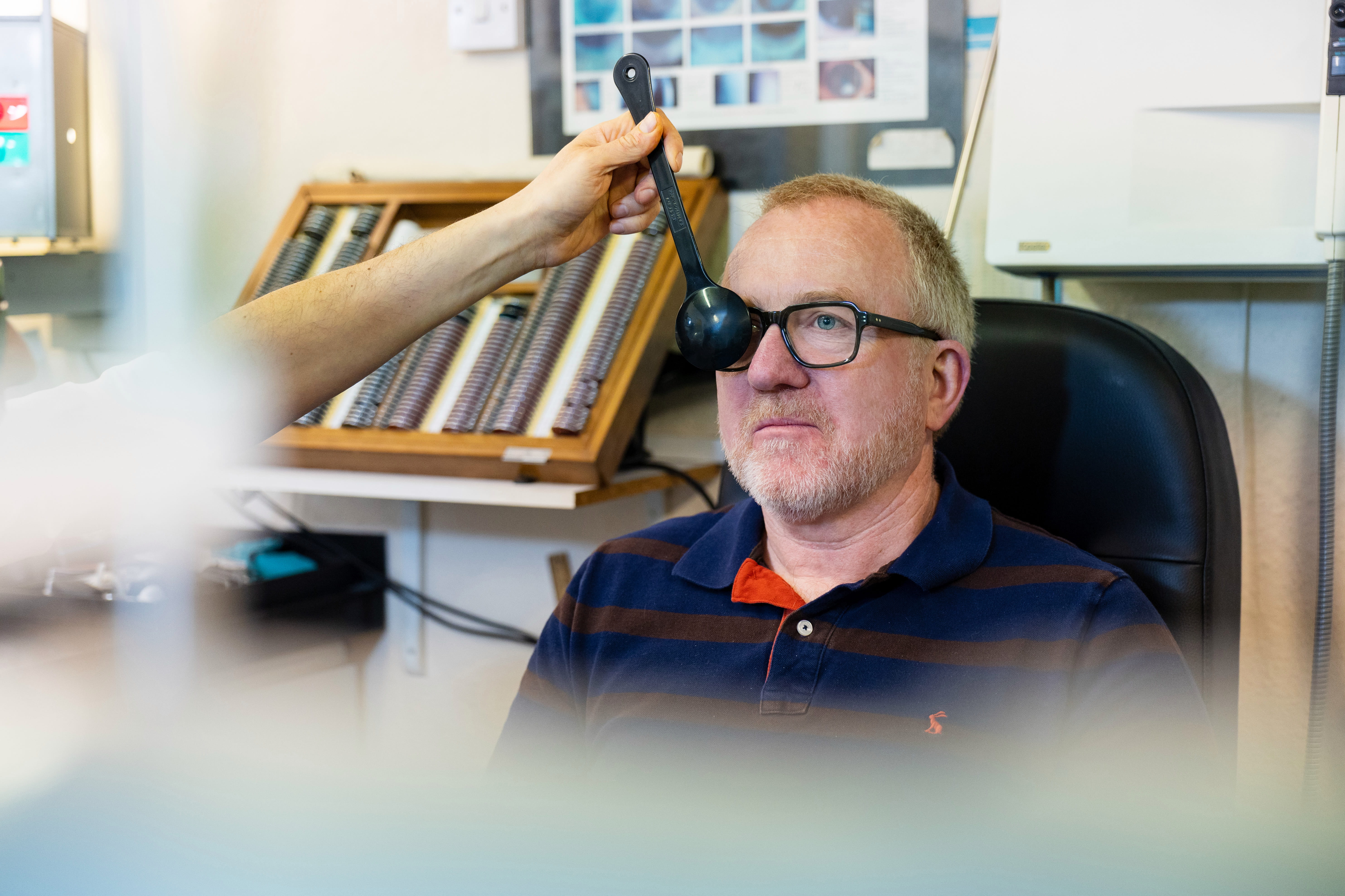 Man wearing glasses having an eye examination with a handheld occluder in an optometrist's office.