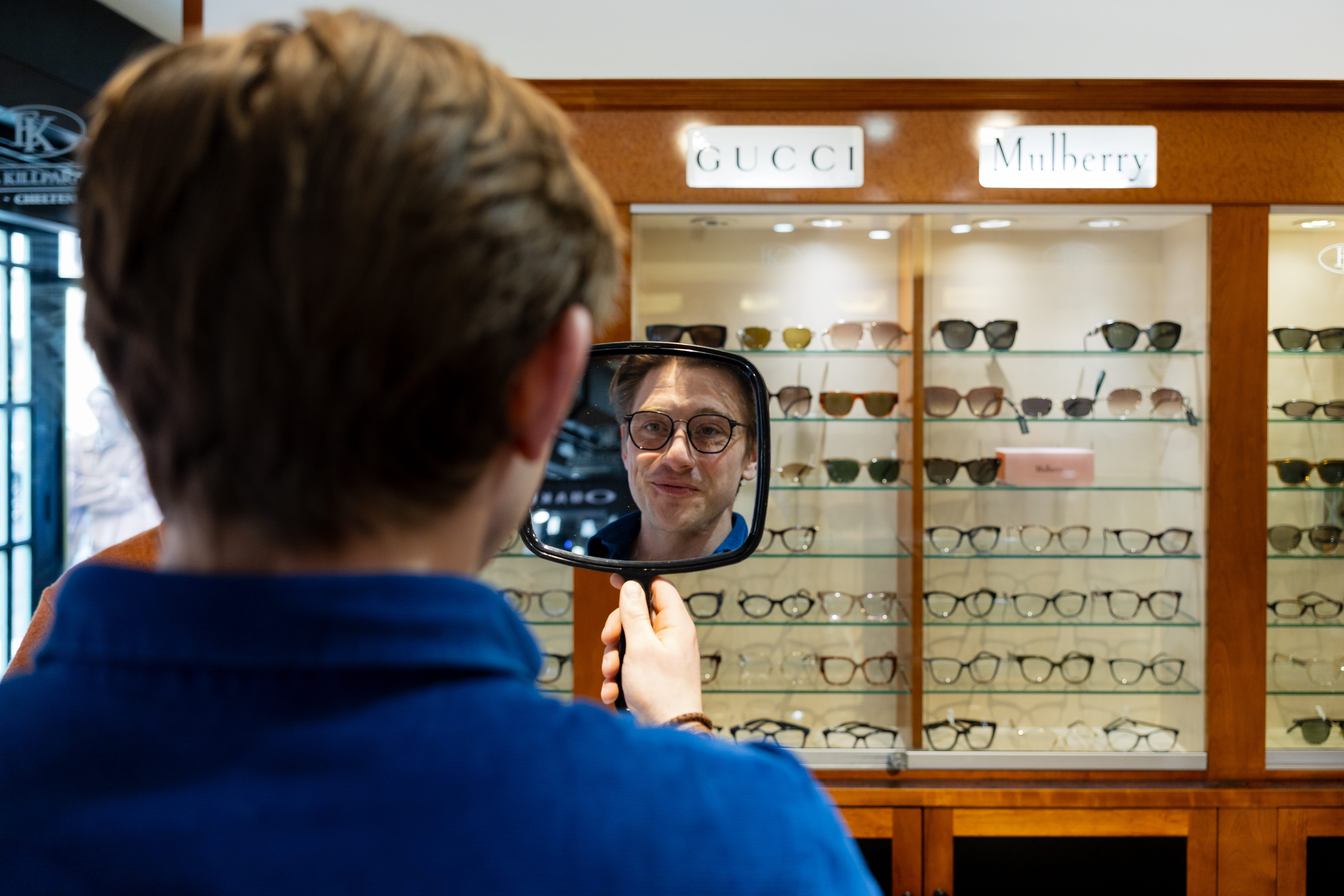Man with glasses holds a mirror showing his reflection in an eyeglasses store with branded frames.