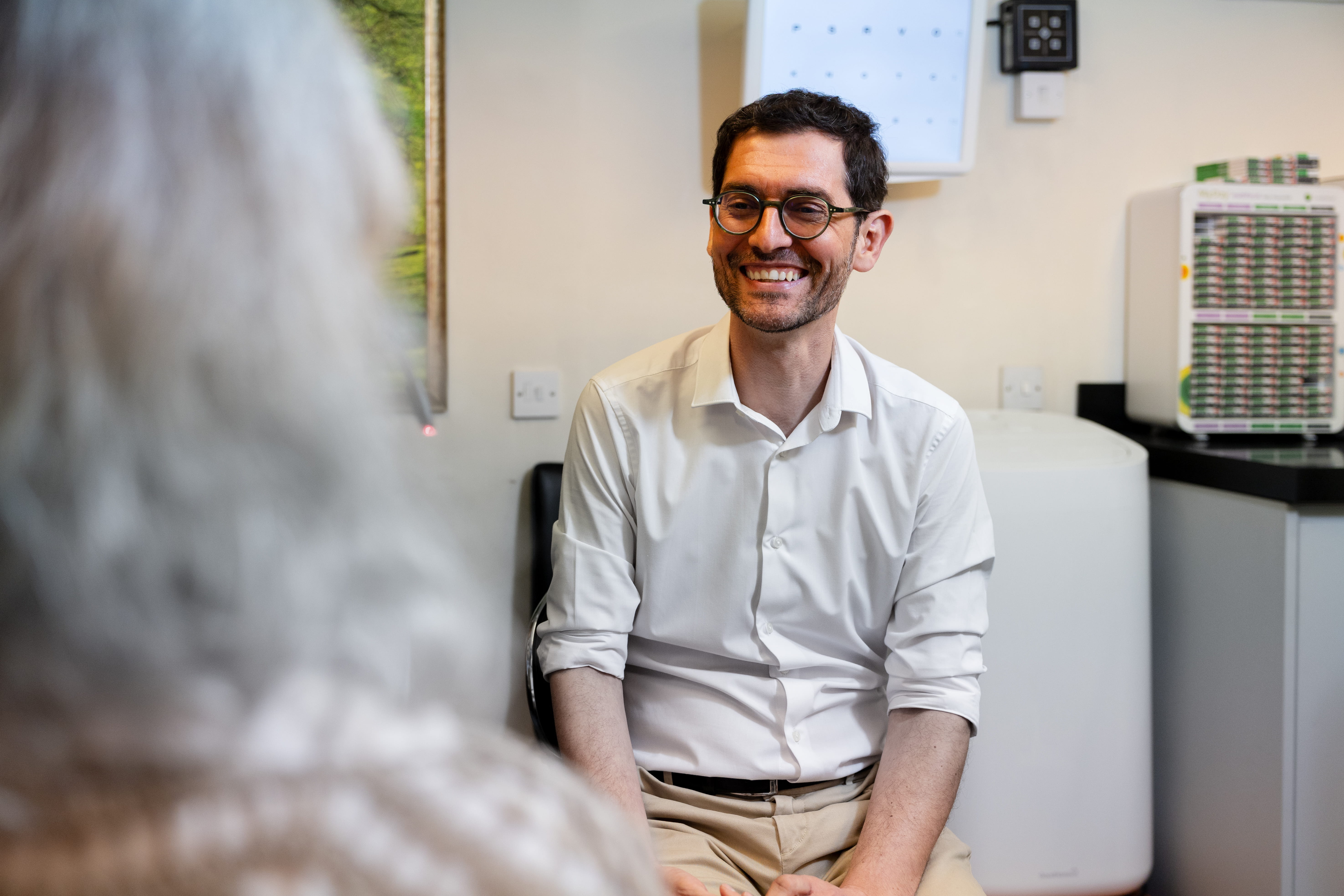 Smiling man with glasses wearing a white shirt, sitting and talking to a person with gray hair.