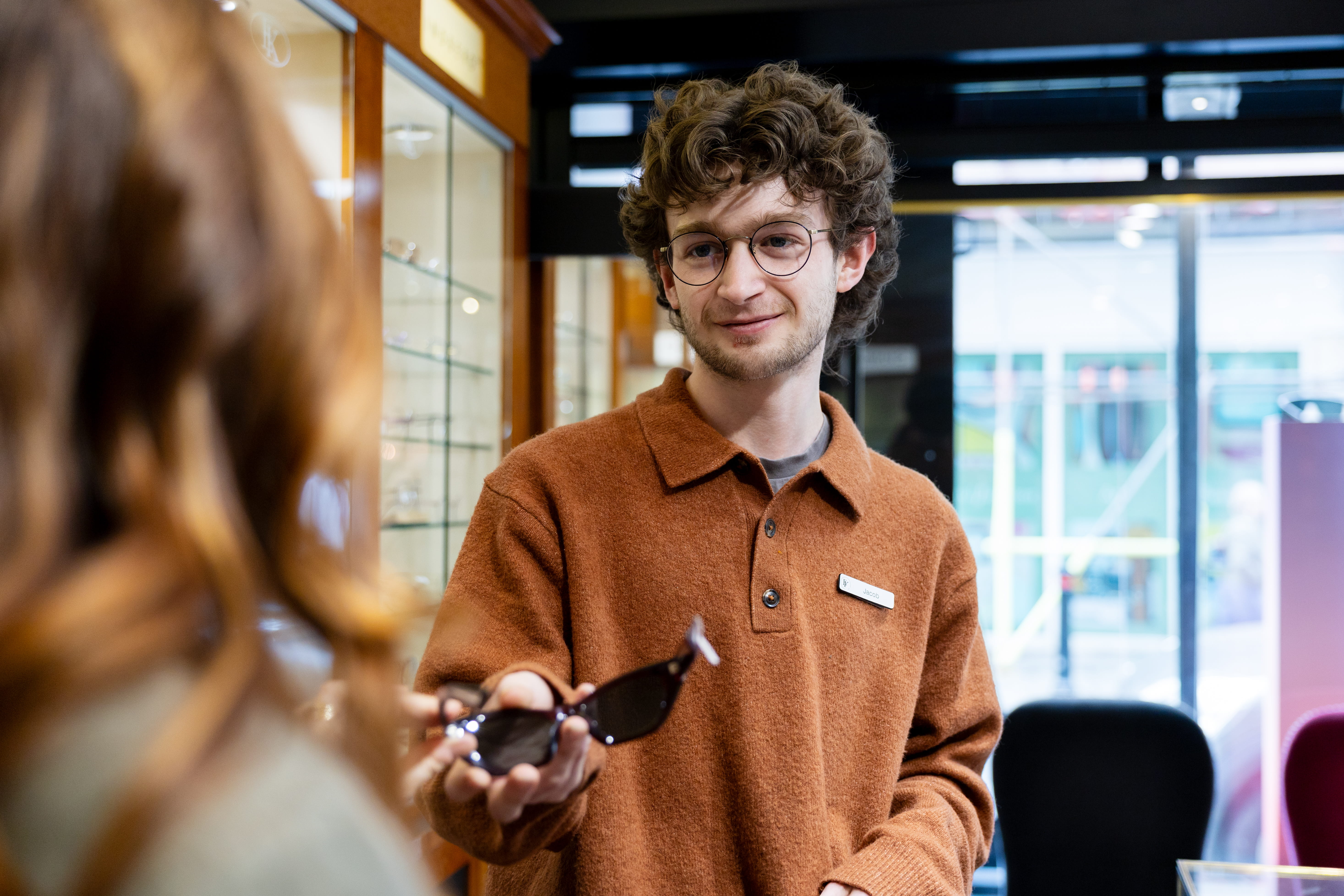 Young man in glasses and brown sweater handing sunglasses to a customer in an eyewear shop.