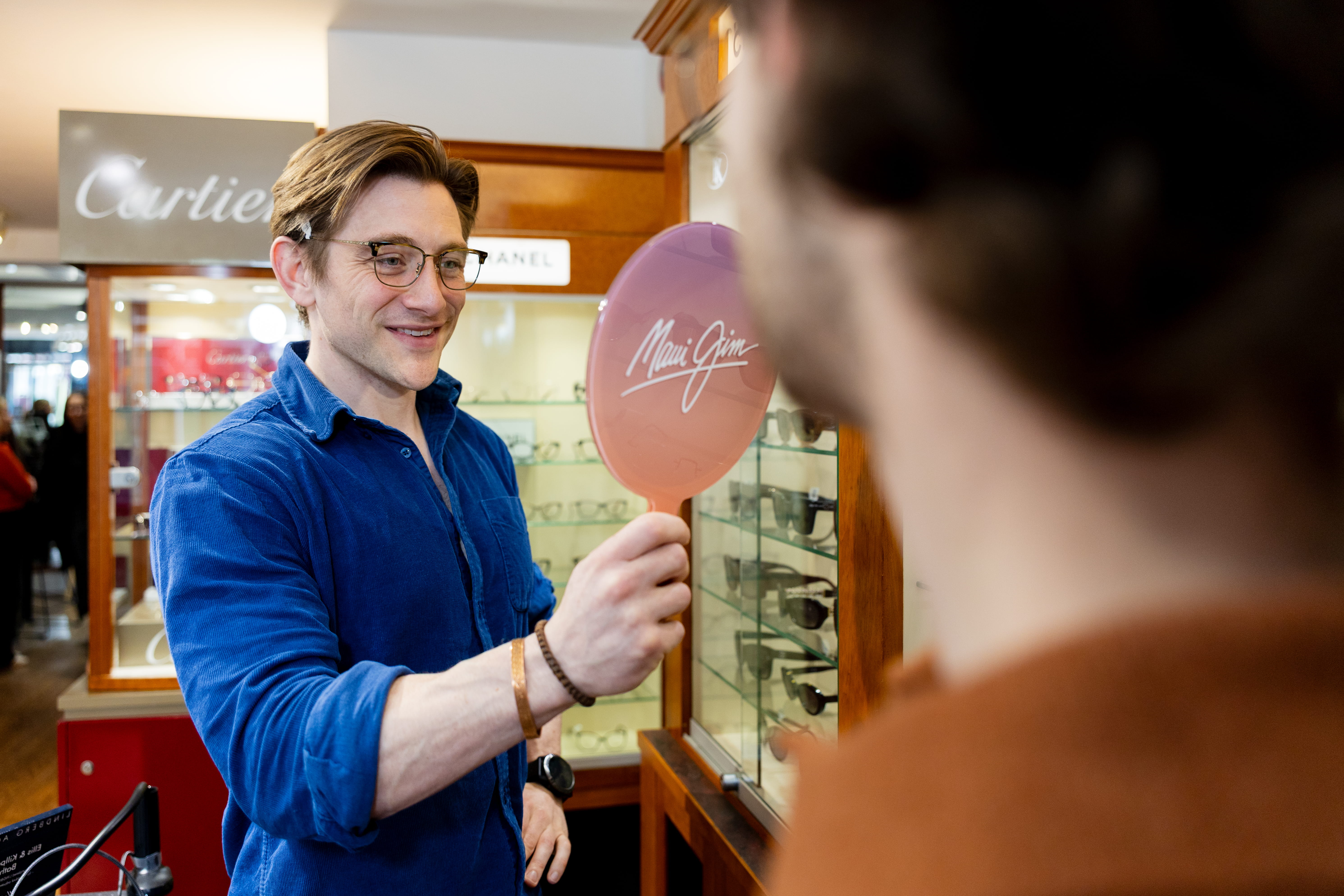 A man in glasses and a blue shirt holds a Maui Jim mirror showing sunglasses to a customer.