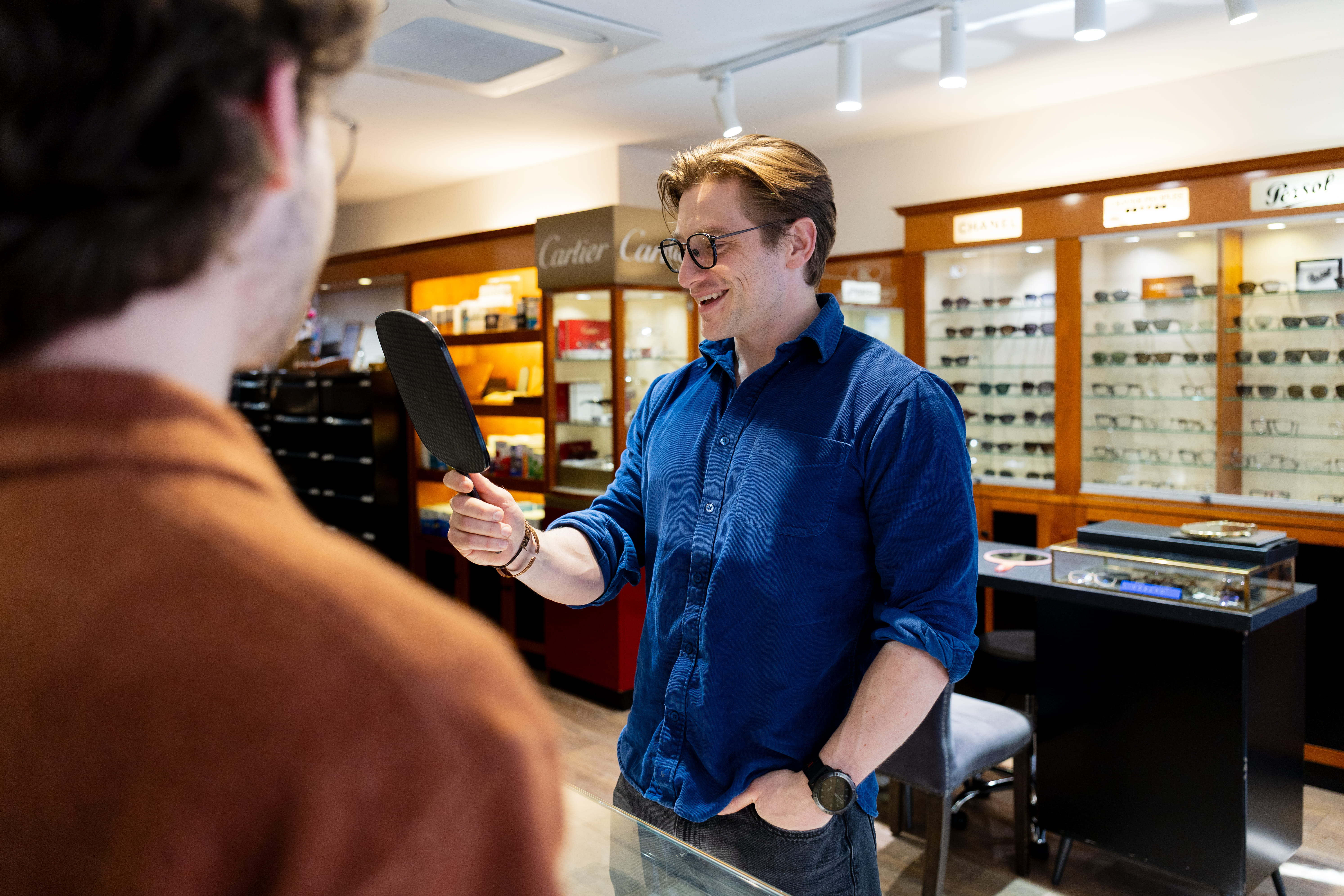 Man in blue shirt holding mirror showing another man his reflection in eyeglass store.