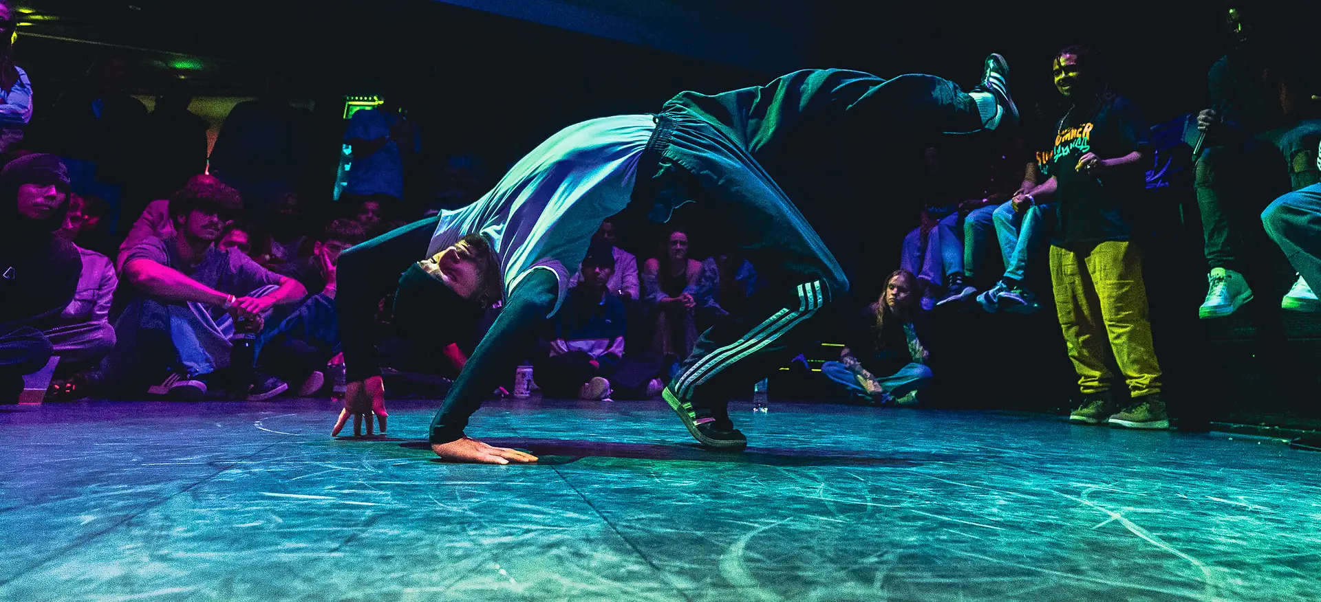 Breakdancer in de Coindrop battle, Vorstin Hilversum, in een brug positie met 1 been in de lucht terwijl mensen er omheen kijken.