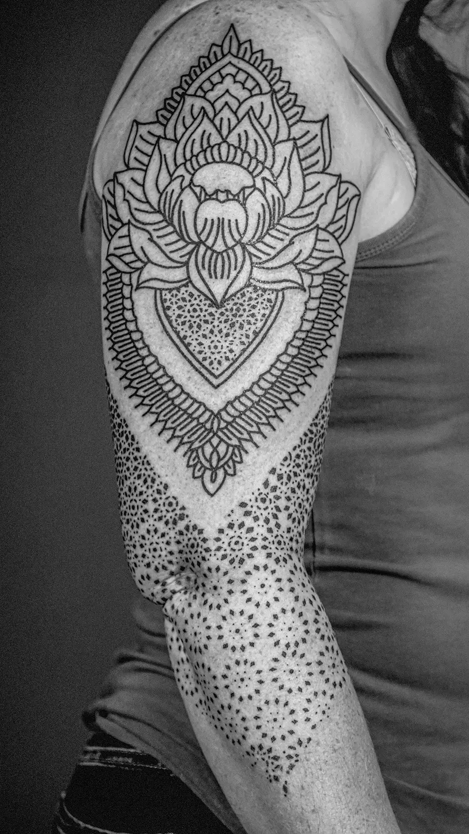 Black and grey tattoo on upper arm featuring a large stylized lotus flower and intricate dotwork patterns extending down the arm.
