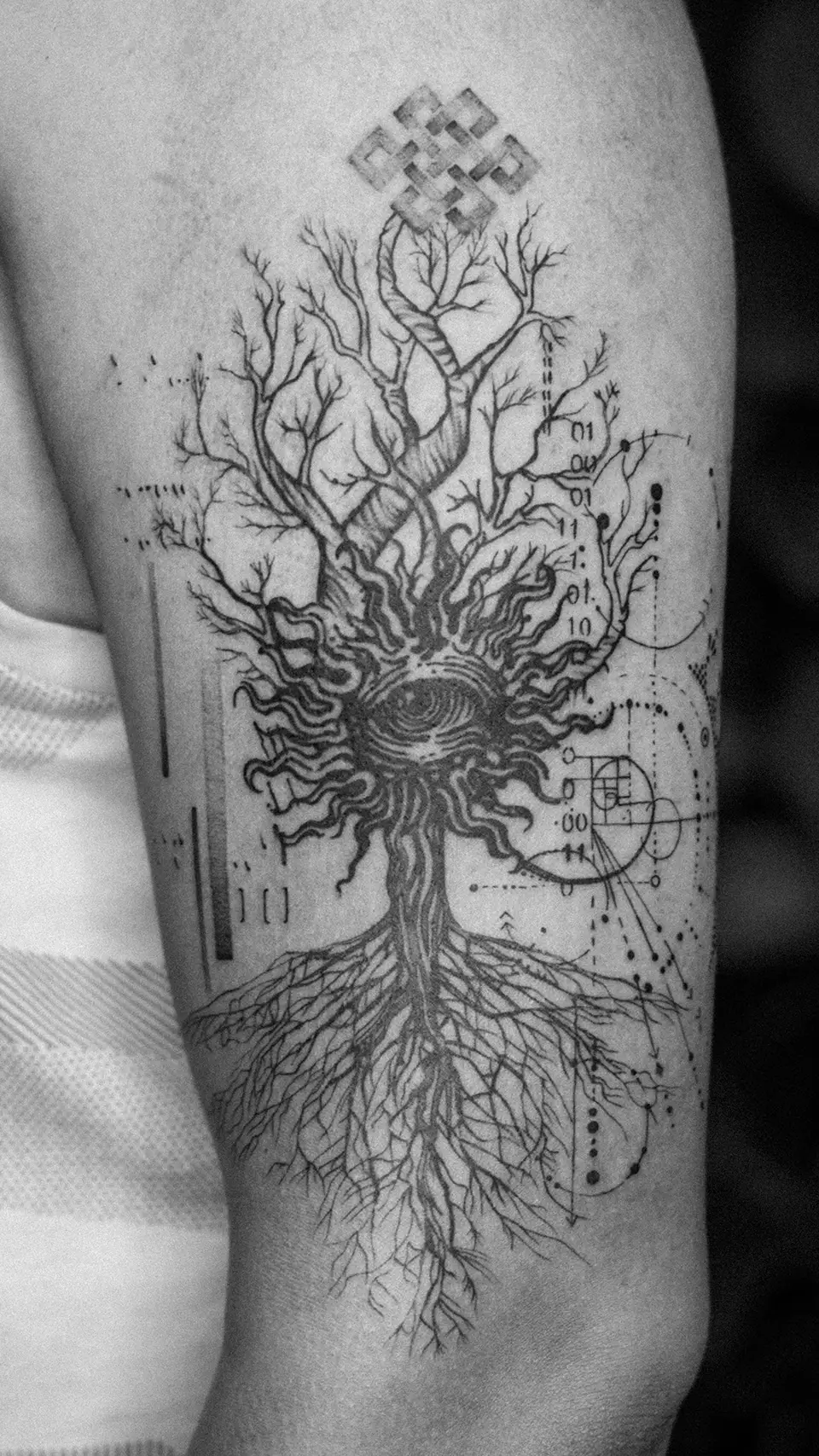 Tattoo of a stylized tree with intricate roots and branches, featuring a circular swirling center and surrounded by abstract geometric designs and binary numbers.
