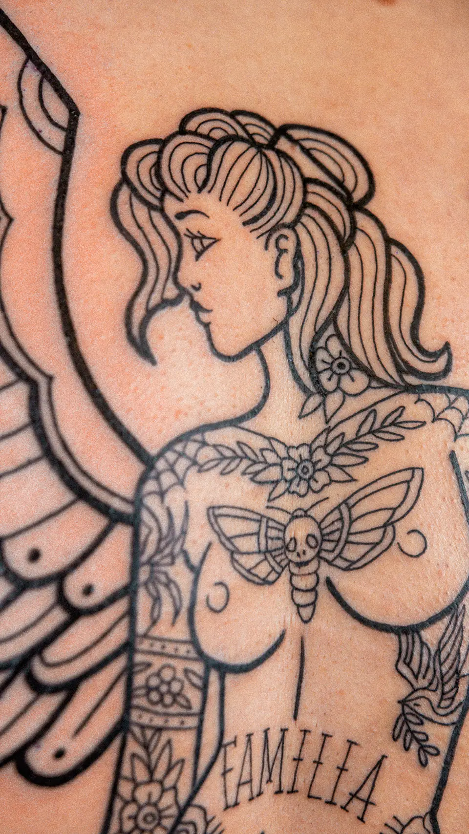 Black ink tattoo of a woman with stylized hair, floral designs, a moth with a skull on its thorax, and the word 'FAMILIA' on her torso.