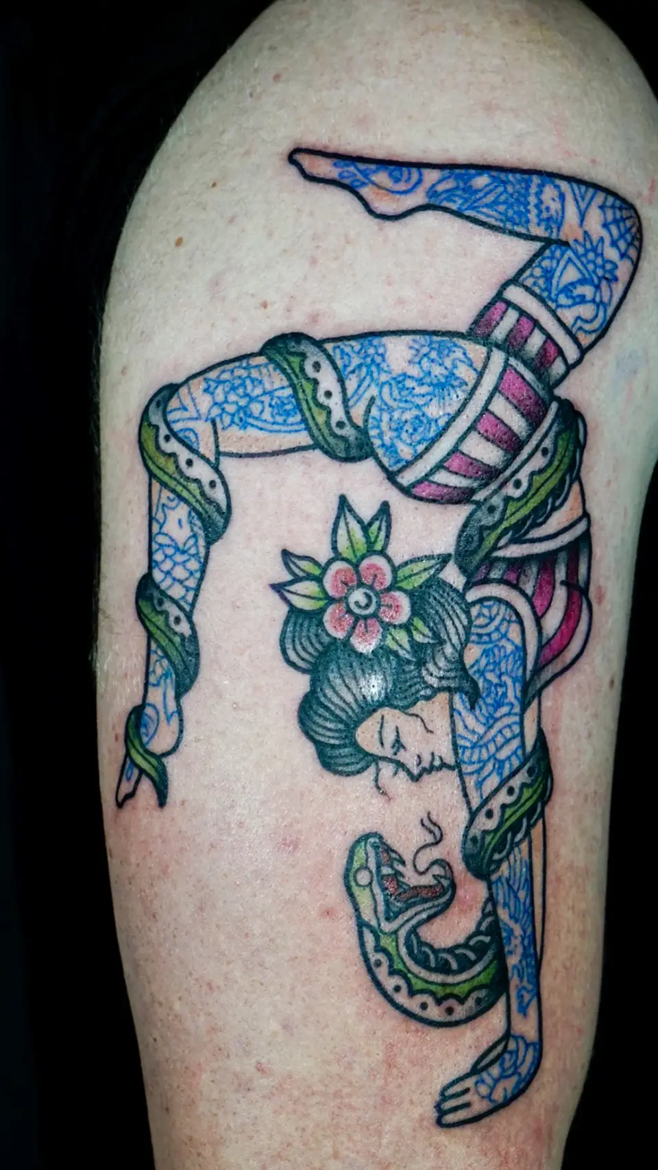 Tattoo of a woman doing a backbend with detailed blue lace patterns and a pink flower in her black hair, entwined with a green snake.