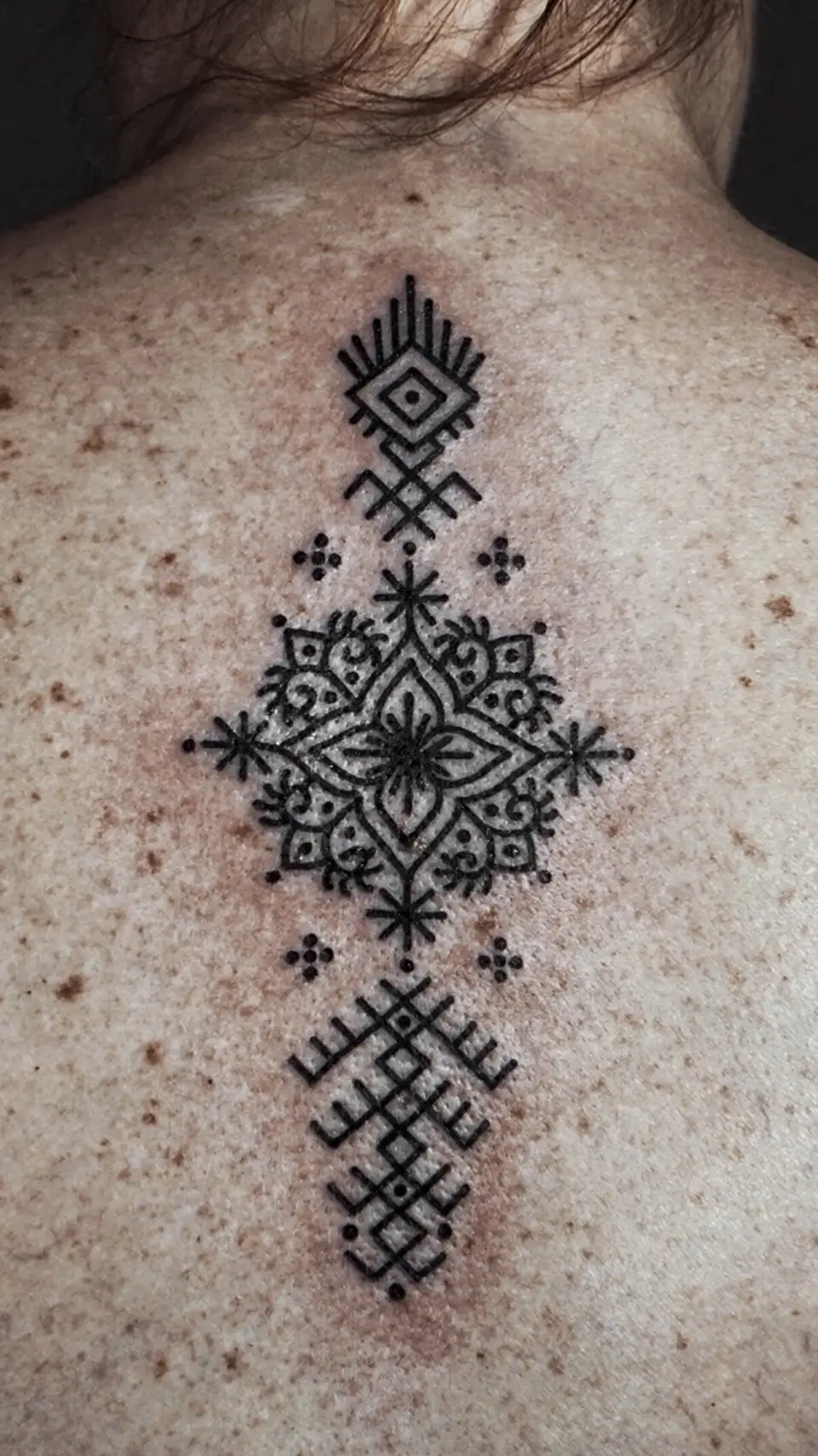 Black geometric and floral tattoo with intricate symmetrical patterns on freckled upper back skin.