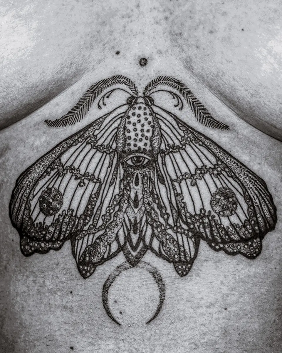 Black ink tattoo of a detailed moth with an eye on its body and crescent moon below.