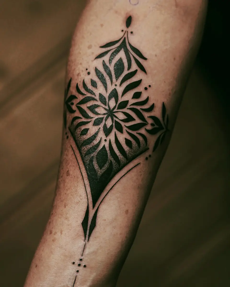 Forearm tattoo of a symmetrical black floral pattern with leaves and dots in a diamond shape.