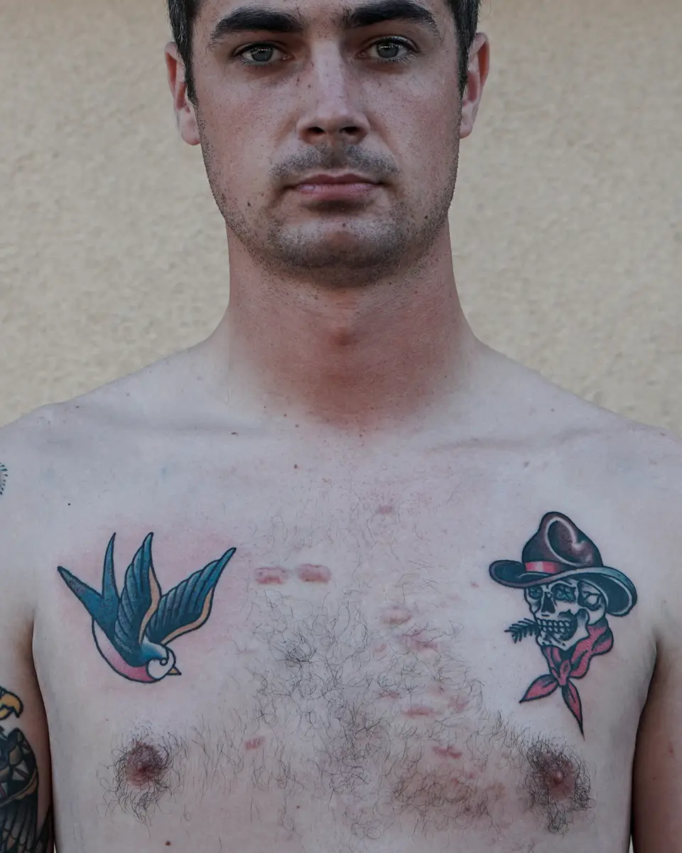 Close-up of a shirtless man with a blue and pink bird tattoo on the left chest and a skull wearing a cowboy hat tattoo on the right chest.