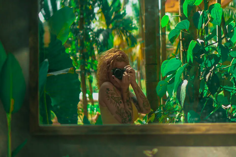 Author of the page with curly hair and tattoos taking a photo with a camera, reflected in a window surrounded by green foliage.