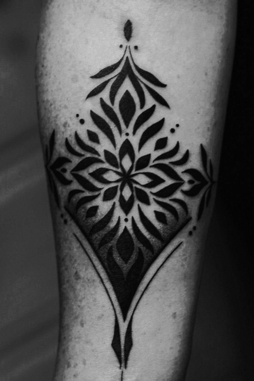 Forearm tattoo of a symmetrical black floral pattern with leaves and dots in a diamond shape.