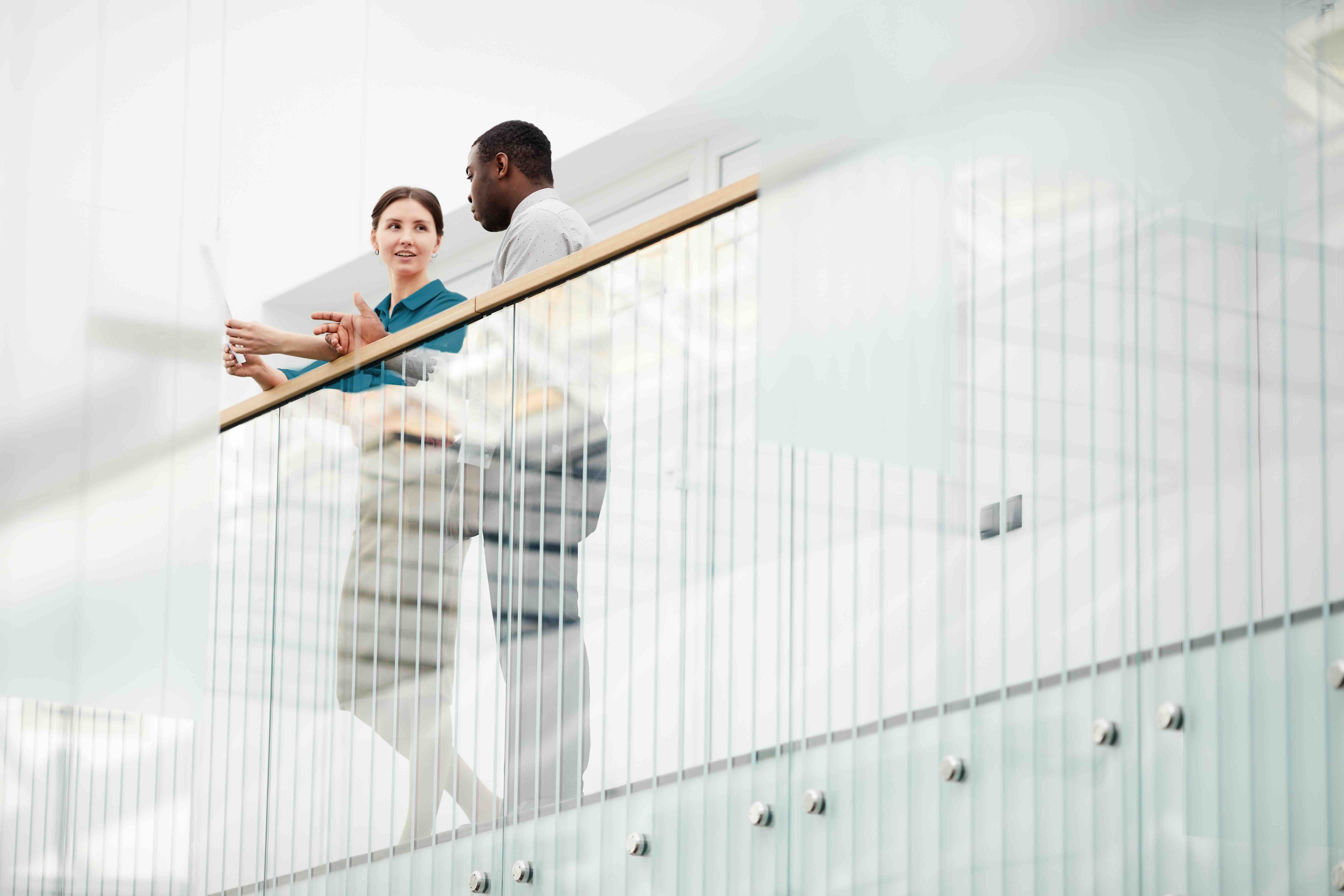 Two colleagues, a woman and a man, standing and discussing while holding papers near a glass railing in a modern office building.