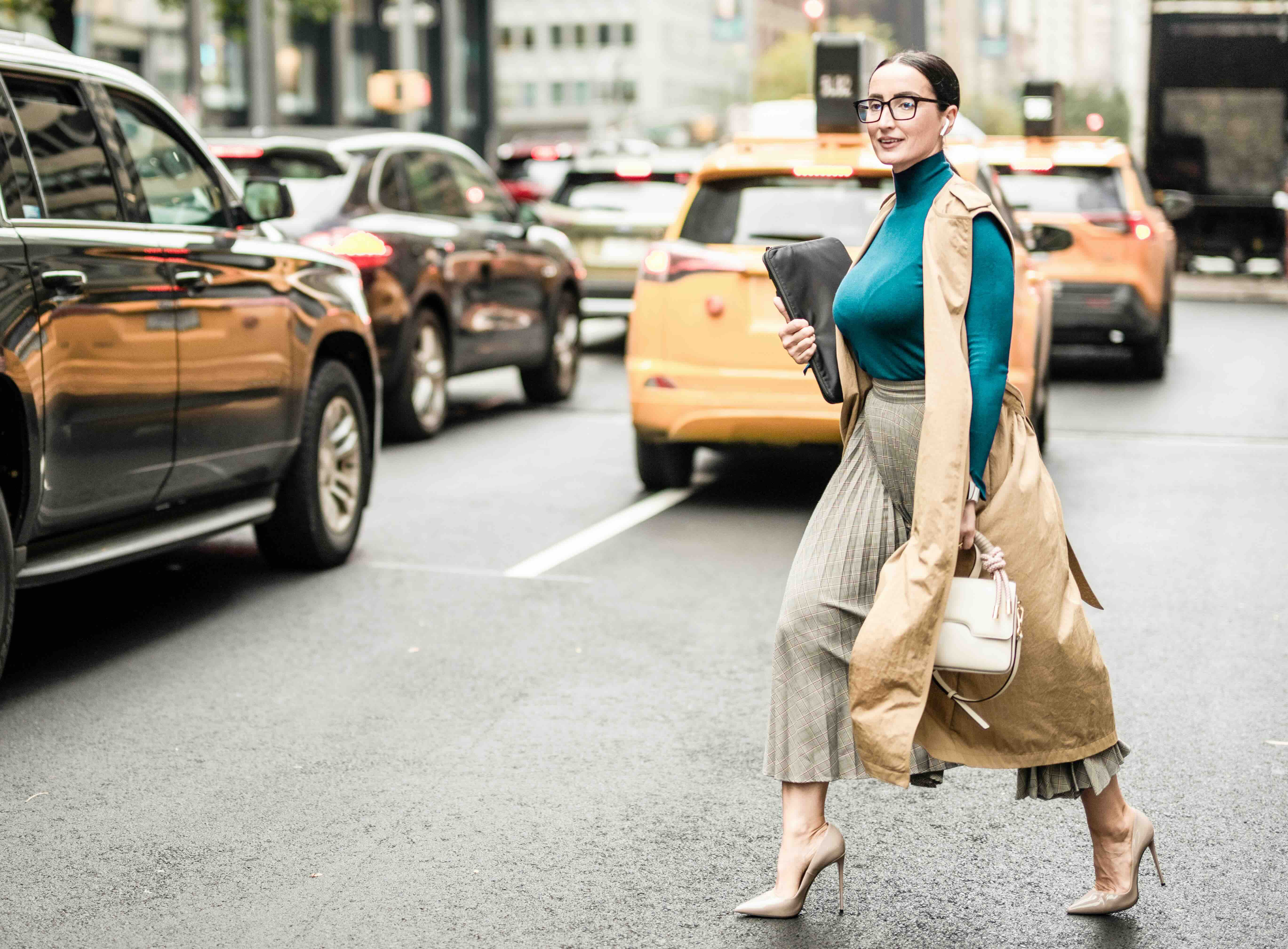 Stylish woman in glasses and trench coat crossing a busy city street with yellow taxis and cars in the background.