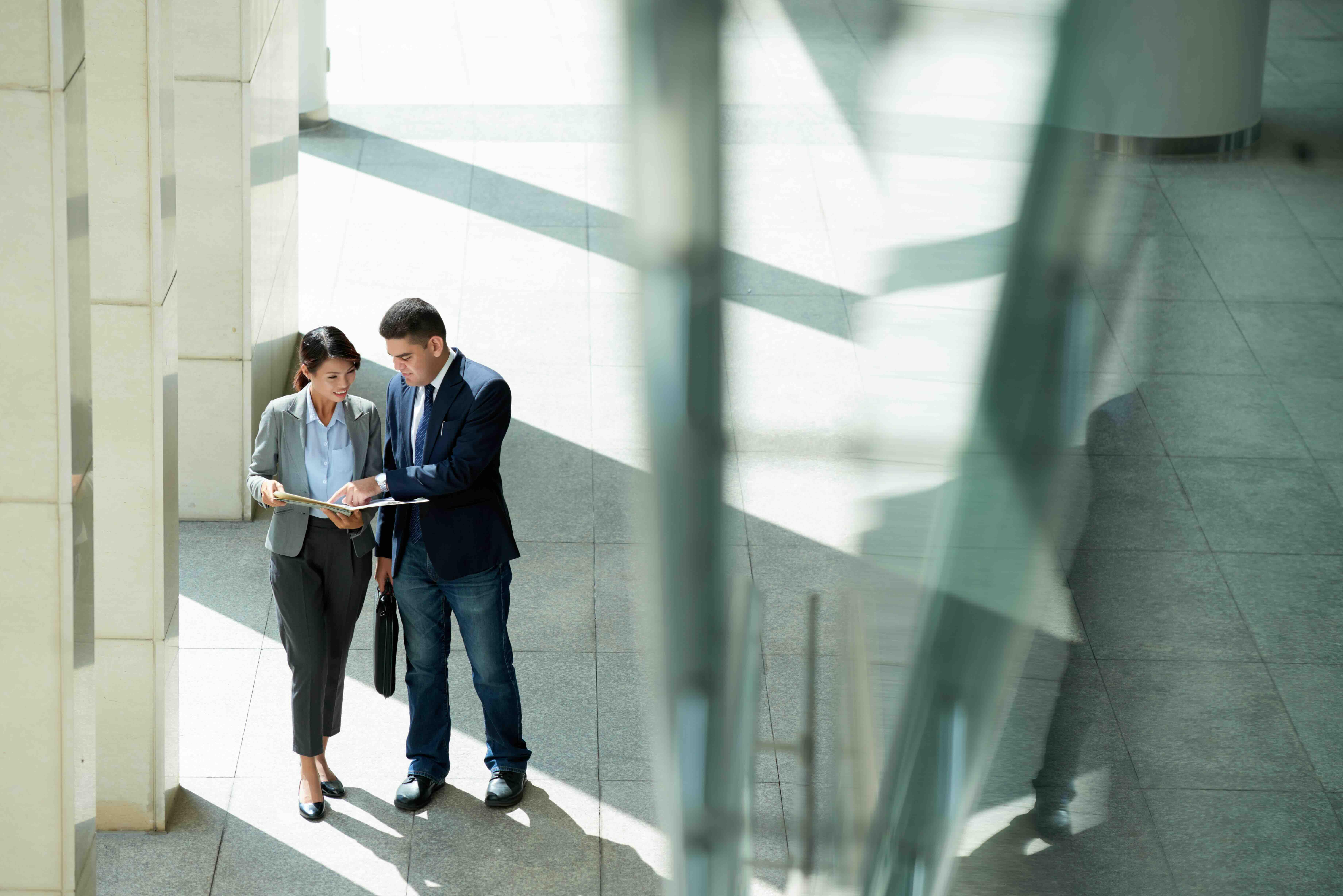 A man and a woman standing together in business attire reviewing a document inside the lobby of a business building with tile floors and lots of light.
