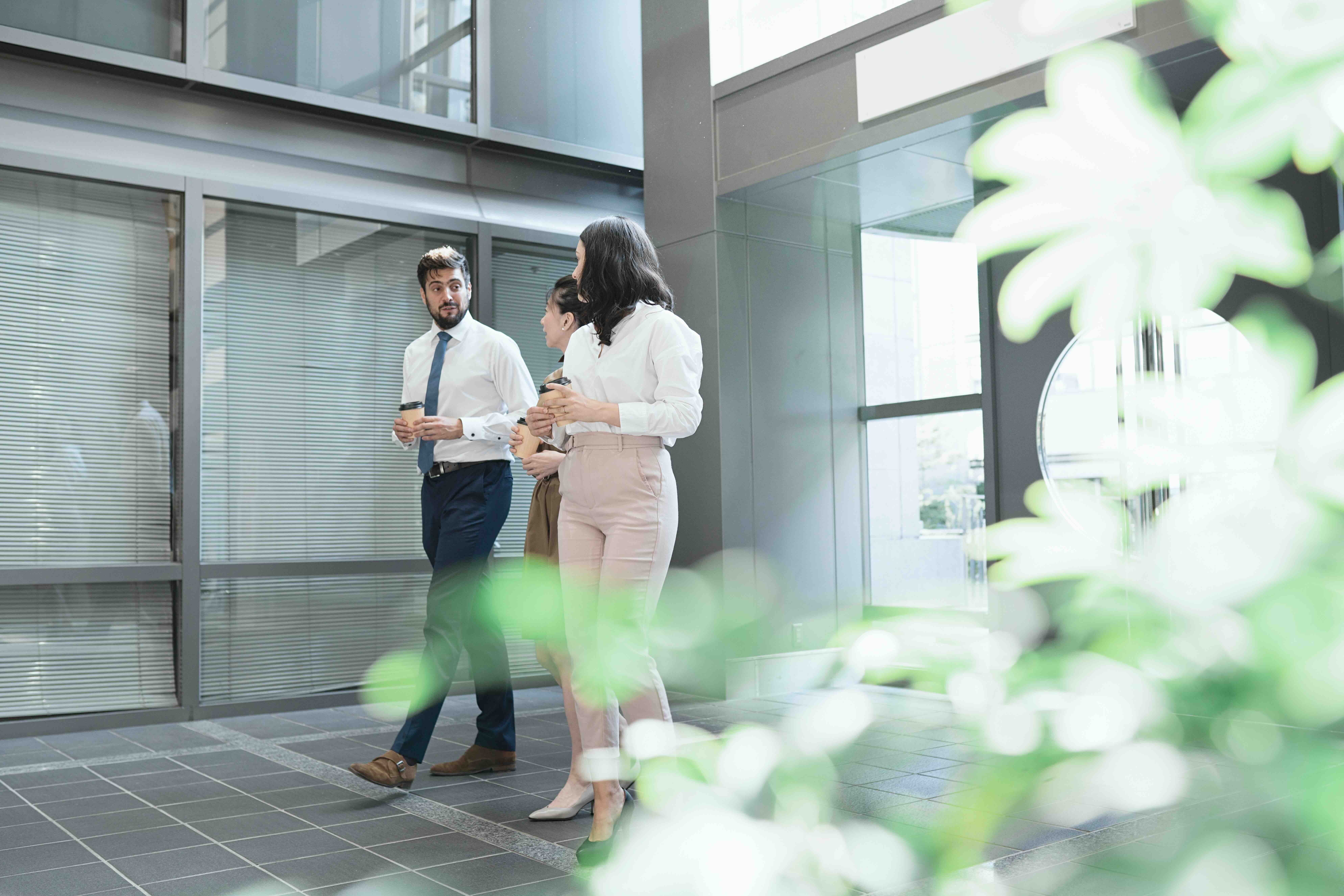 Three colleagues, a man and two women, walking into the lobby of an office building with coffee cups and a plant in the foreground.