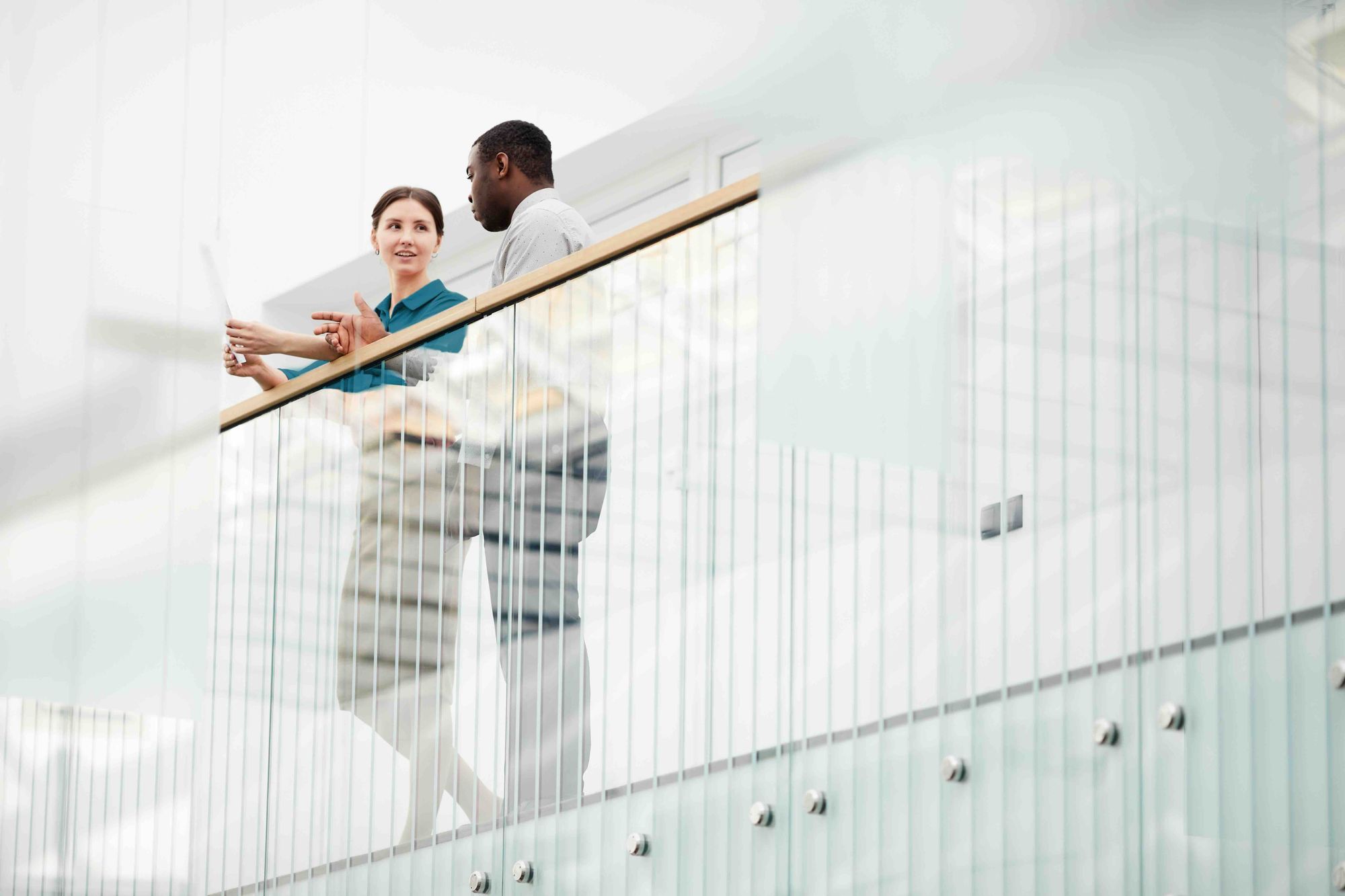 Two colleagues, a woman and a man, standing and discussing while holding papers near a glass railing in a modern office building.