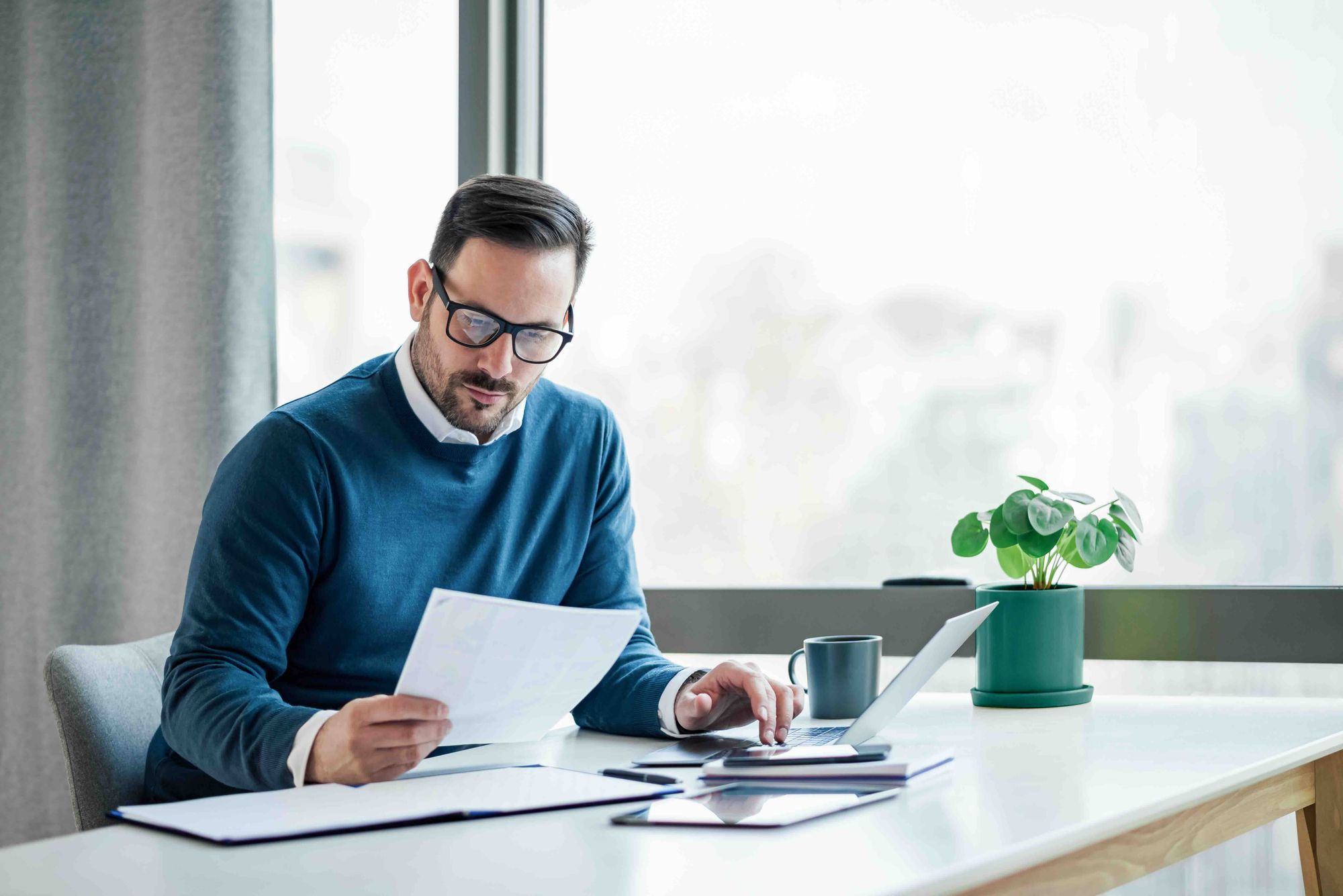 Man wearing glasses and a blue sweater working at a desk with documents, laptop, tablet, and a coffee mug near a window.