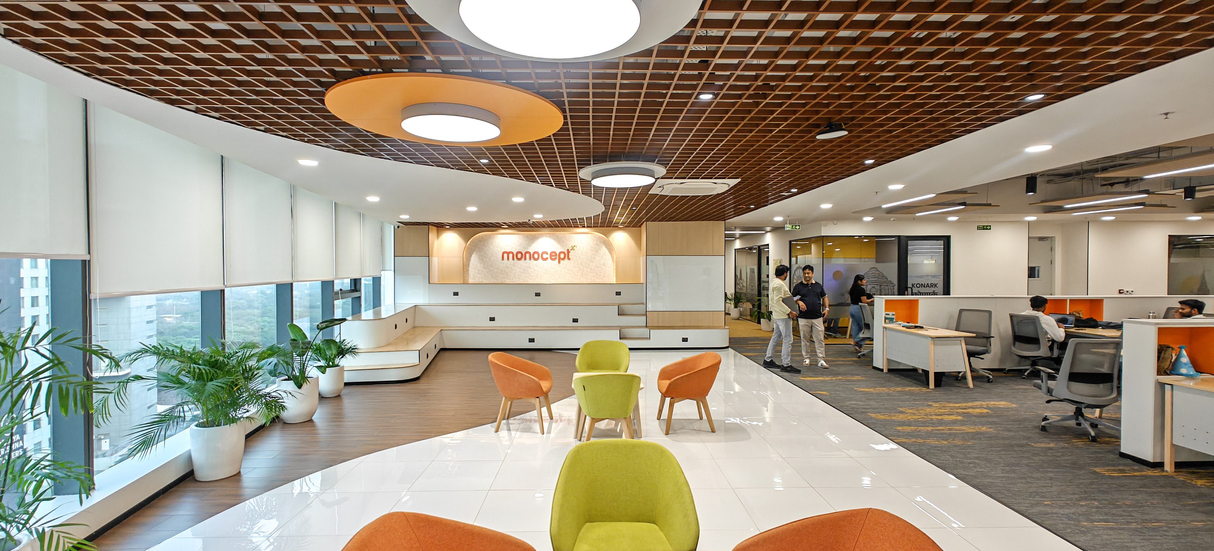 Modern office lounge with orange and green chairs, large windows with blinds, potted plants, and a wooden ceiling grid.