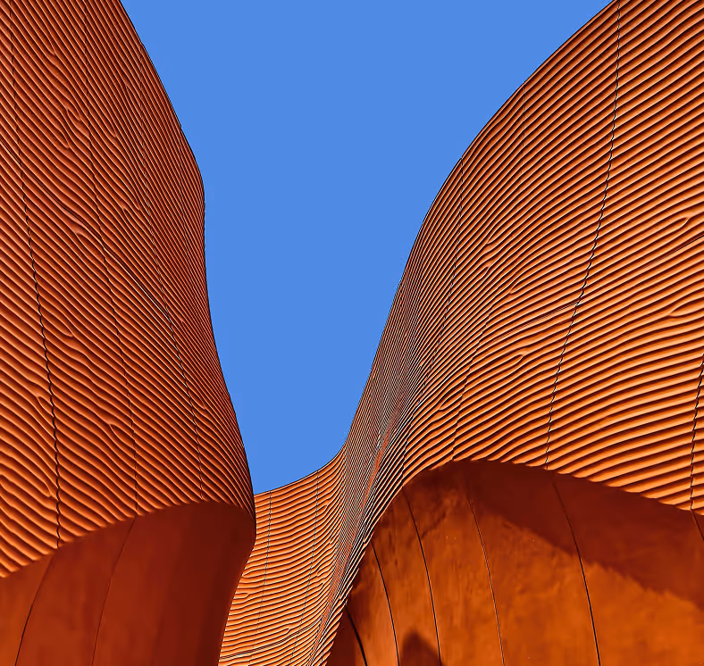 Upward view of curved, orange corrugated metal building walls against a clear blue sky.