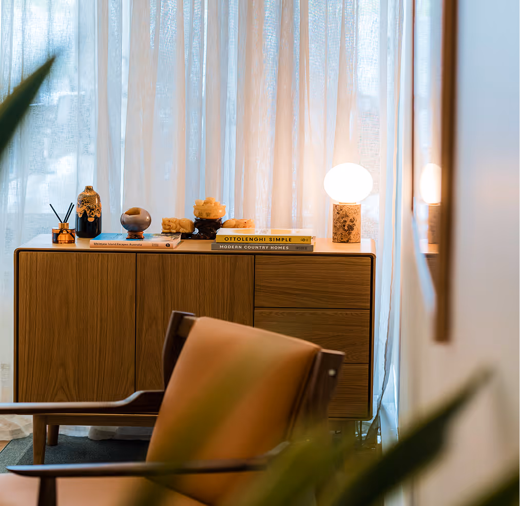 Modern wooden sideboard with decorative items and a glowing round lamp, in front of sheer curtains, with a blurred chair in the foreground.