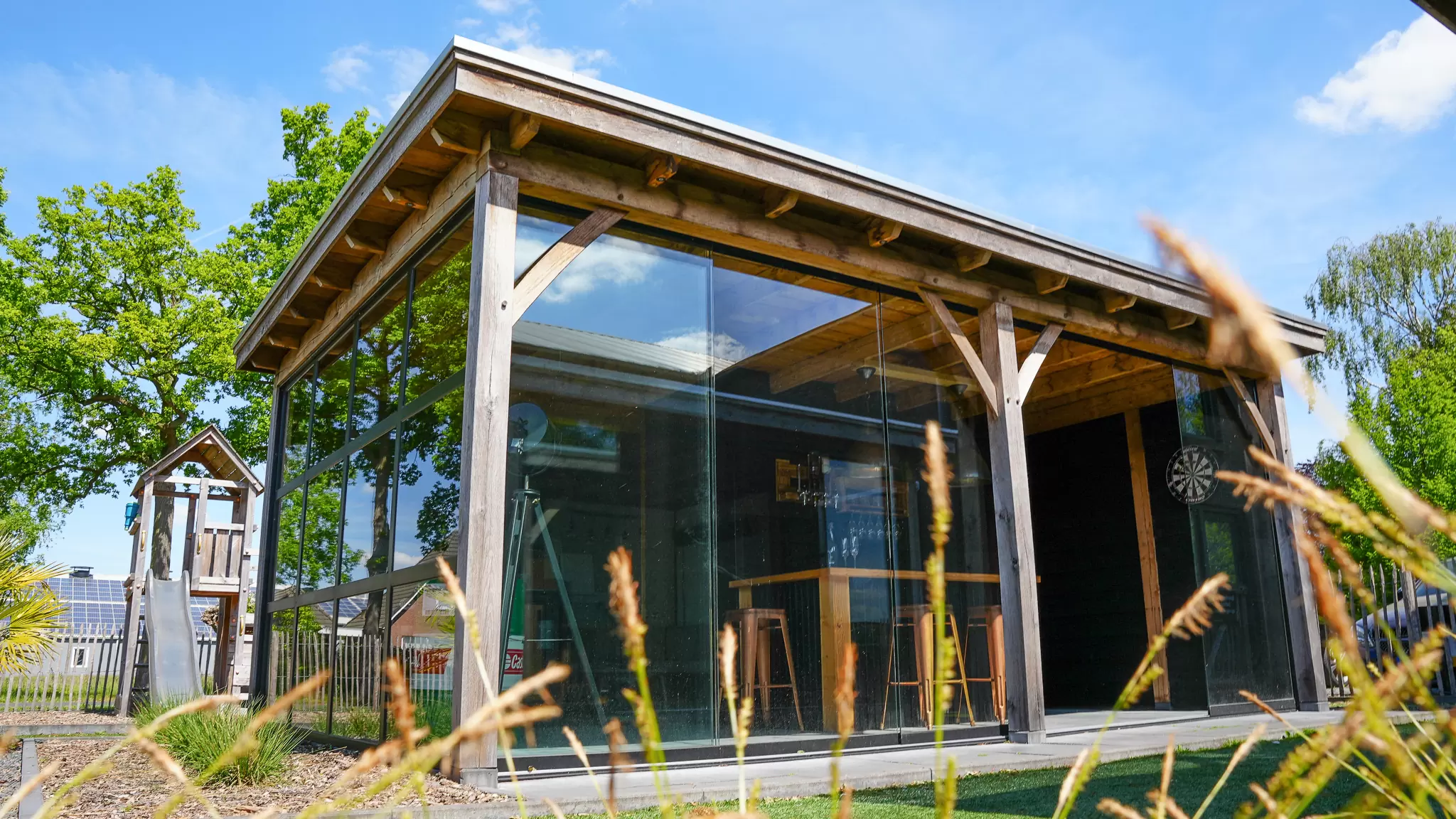 Modern wooden outdoor pavilion with glass walls and wooden bar stools inside, next to a playground slide under a clear blue sky.