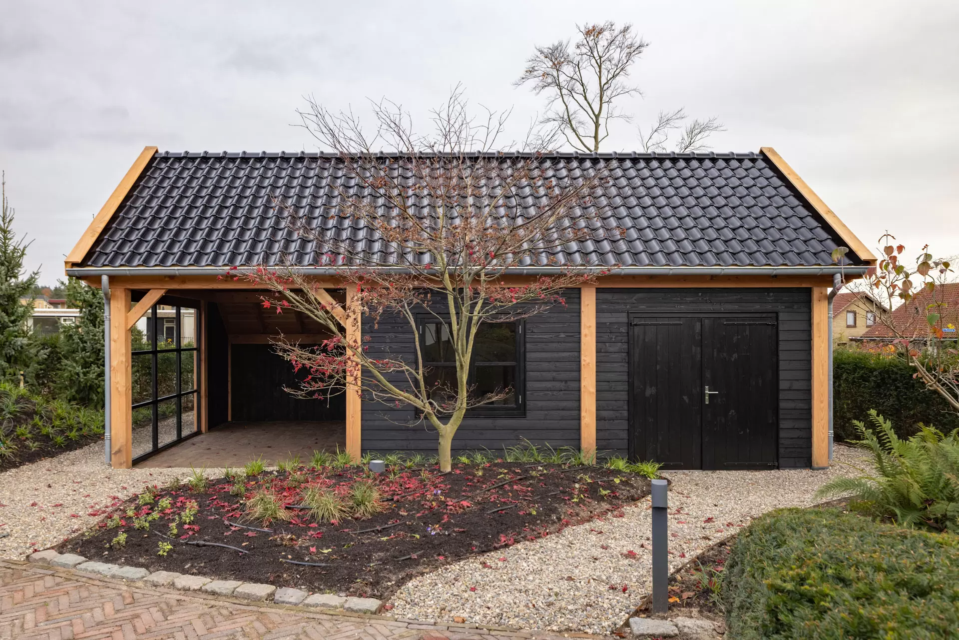 Modern black wooden garden shed with a tiled roof, featuring an open carport on the left and double doors on the right, surrounded by a landscaped garden with a leaf-shedding tree in front.