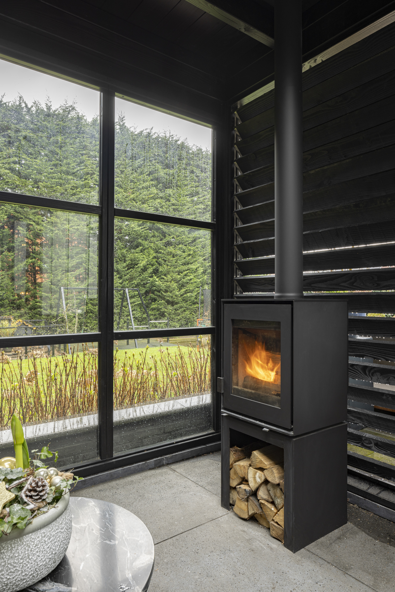 Black wood stove with fire inside next to large window overlooking green garden and trees.