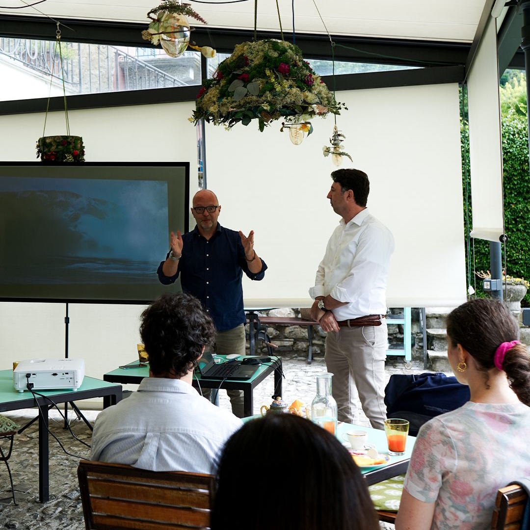 Two men giving a presentation to an audience seated at tables with drinks and snacks, in a room with hanging plants and a projector screen.