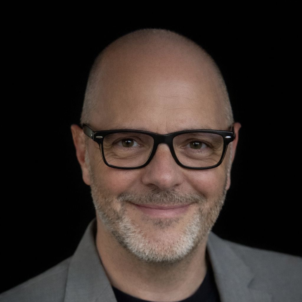 Smiling bald man with glasses and gray beard wearing a gray jacket against a black background.