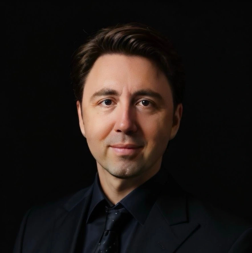 Close-up portrait of a man with light skin and brown hair wearing a white collared shirt against a black background.