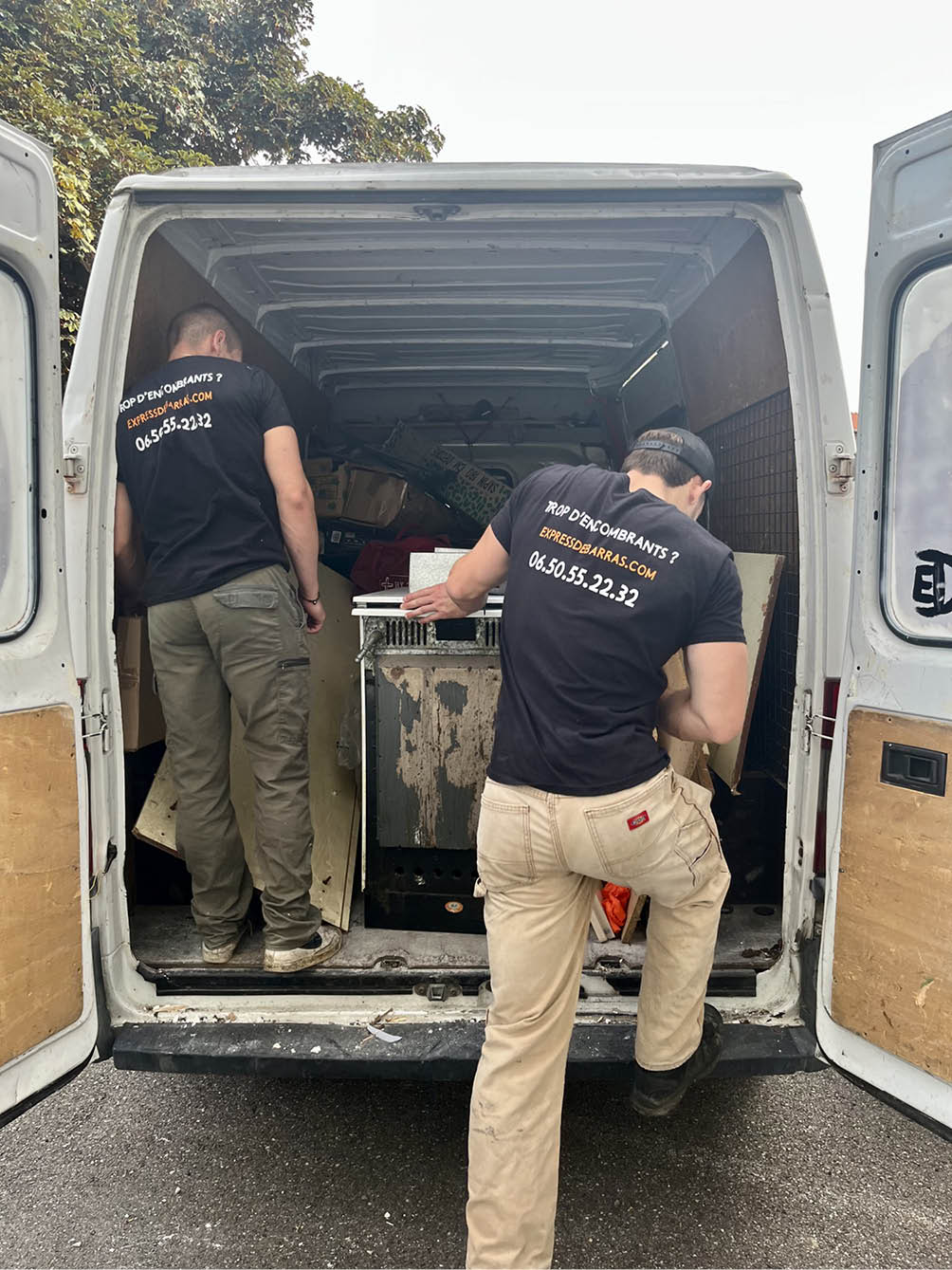 Two men wearing black shirts with removal service contact information loading items into the back of a white van with open doors.